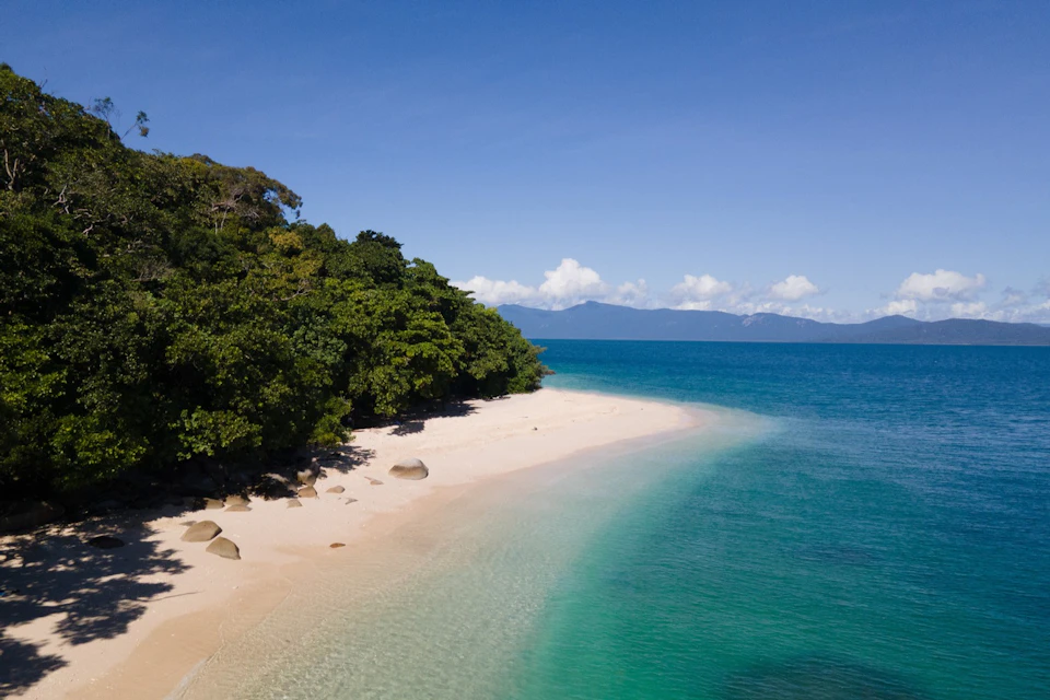 A secluded beach with white sand, clear turquoise water, large boulders, and lush green trees under a clear blue sky.