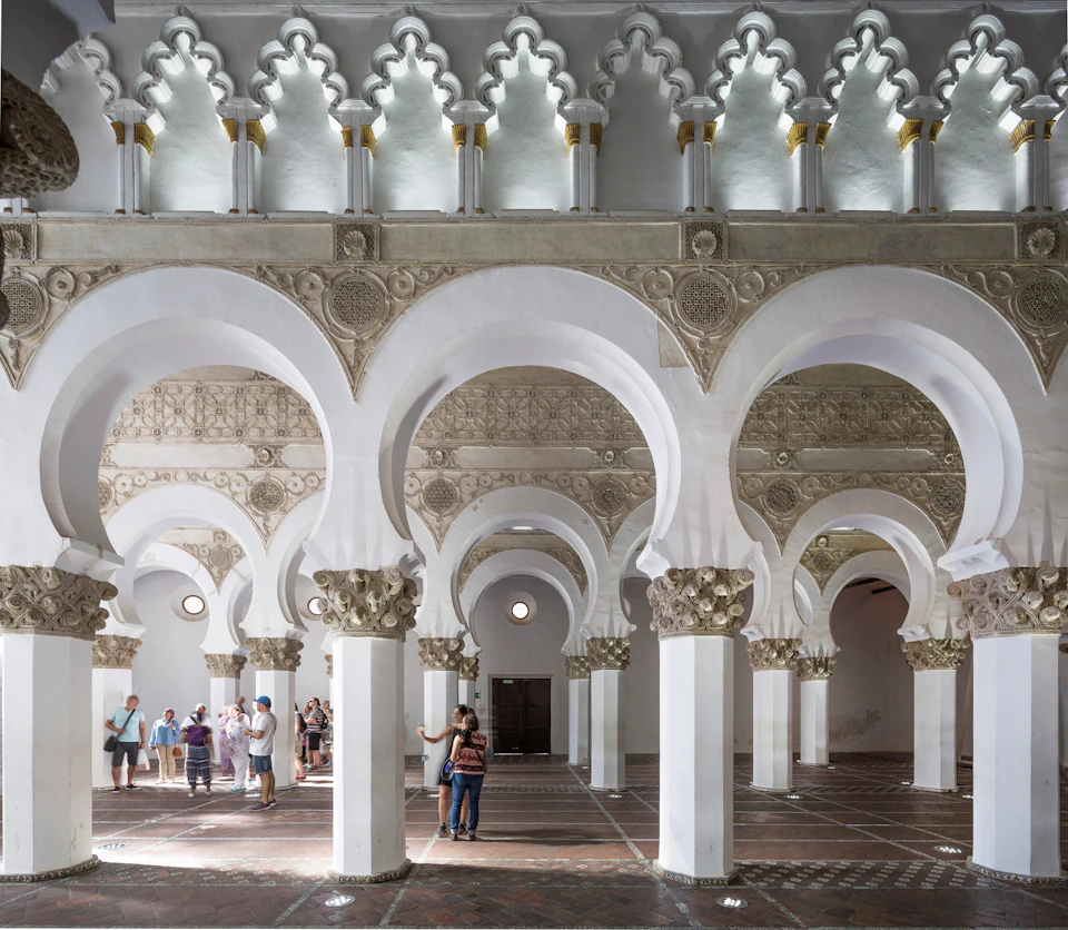 People walk through a historic hall filled with elaborately carved arches, columns, and intricate designs on the walls and ceiling.