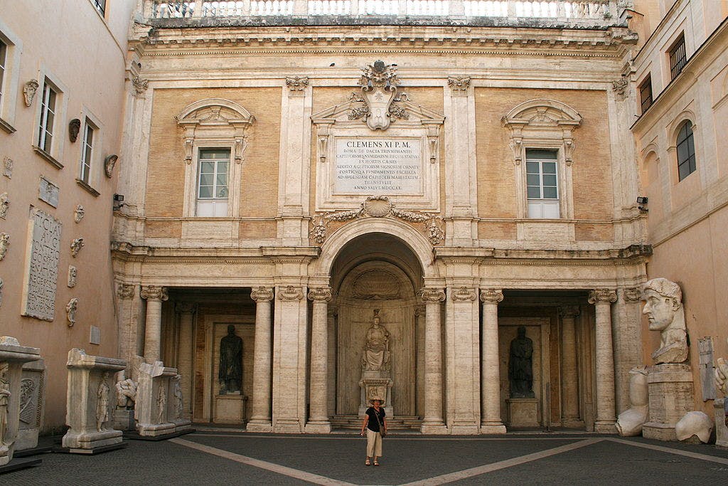 Person stands in front of a historic building facade with columns, statues, and arched entrances. Text is engraved above.
