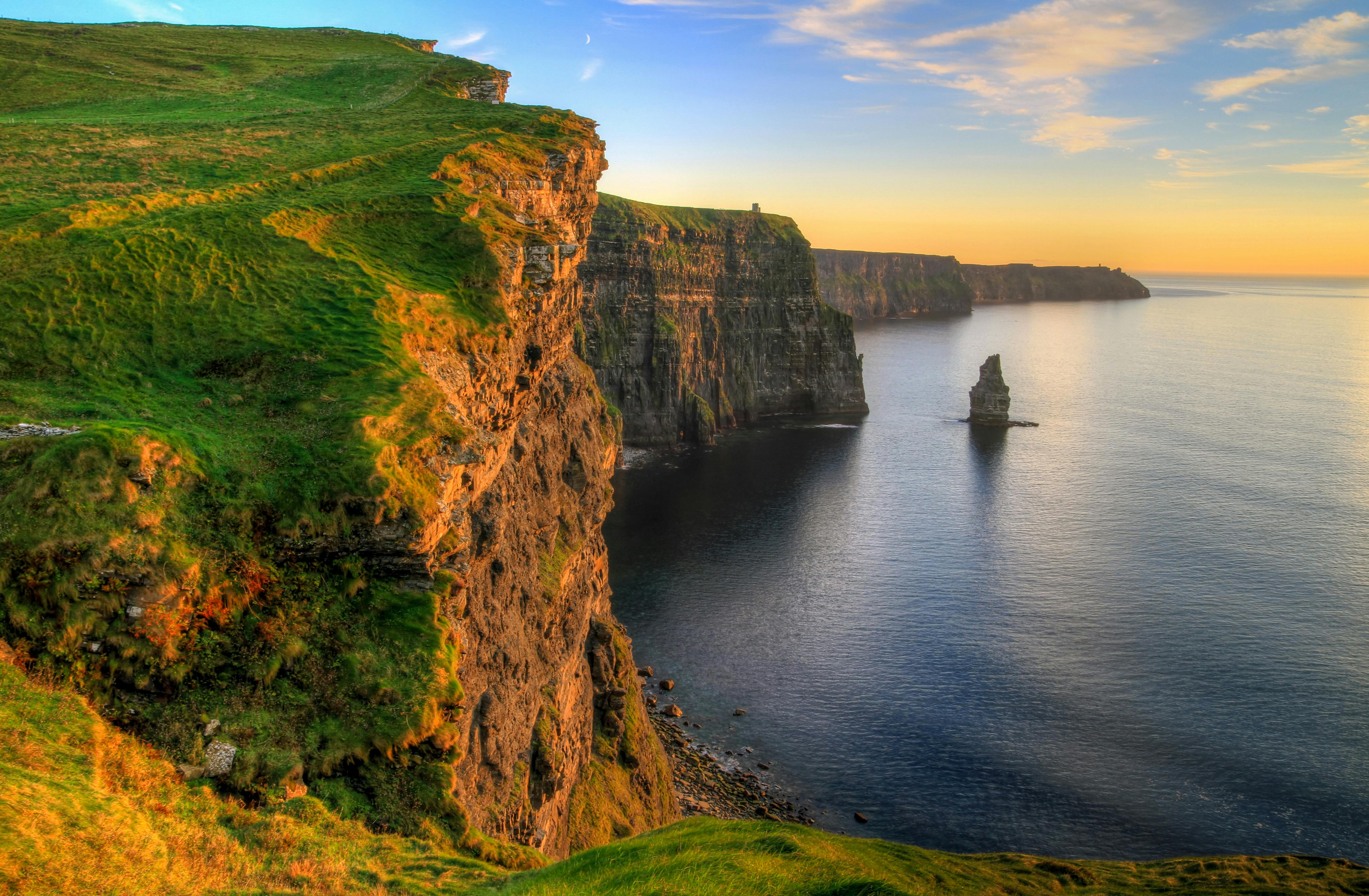 Steep cliffs covered in green grass overlooking calm ocean waters at sunrise, with a sea stack near the shore.
