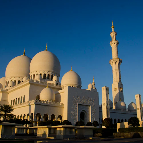 Eine weiße Moschee mit großen Kuppeln und hohen Minaretten unter einem strahlend blauen Himmel, umgeben von gepflegten Grünanlagen und Spazierwegen.