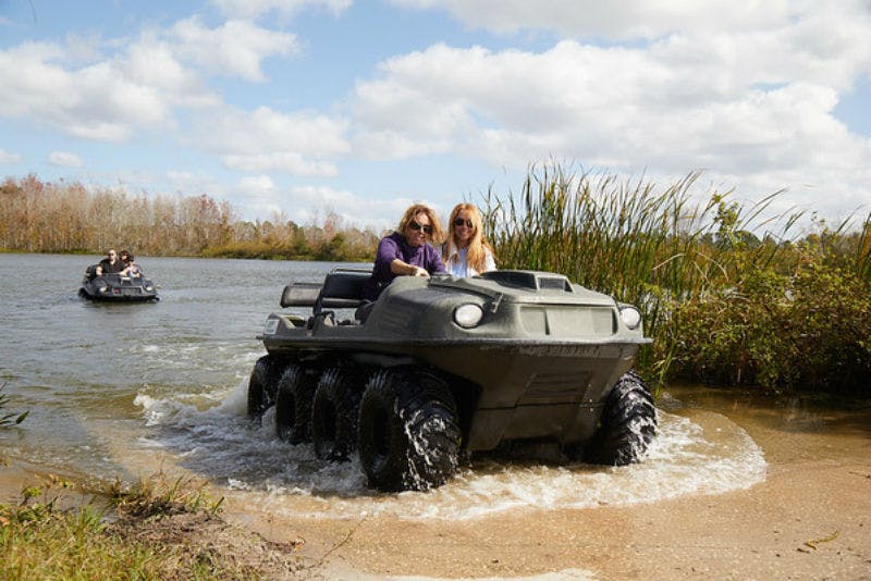 Two people riding a six-wheeled amphibious vehicle, exiting water onto grassy shore with another vehicle in the background.