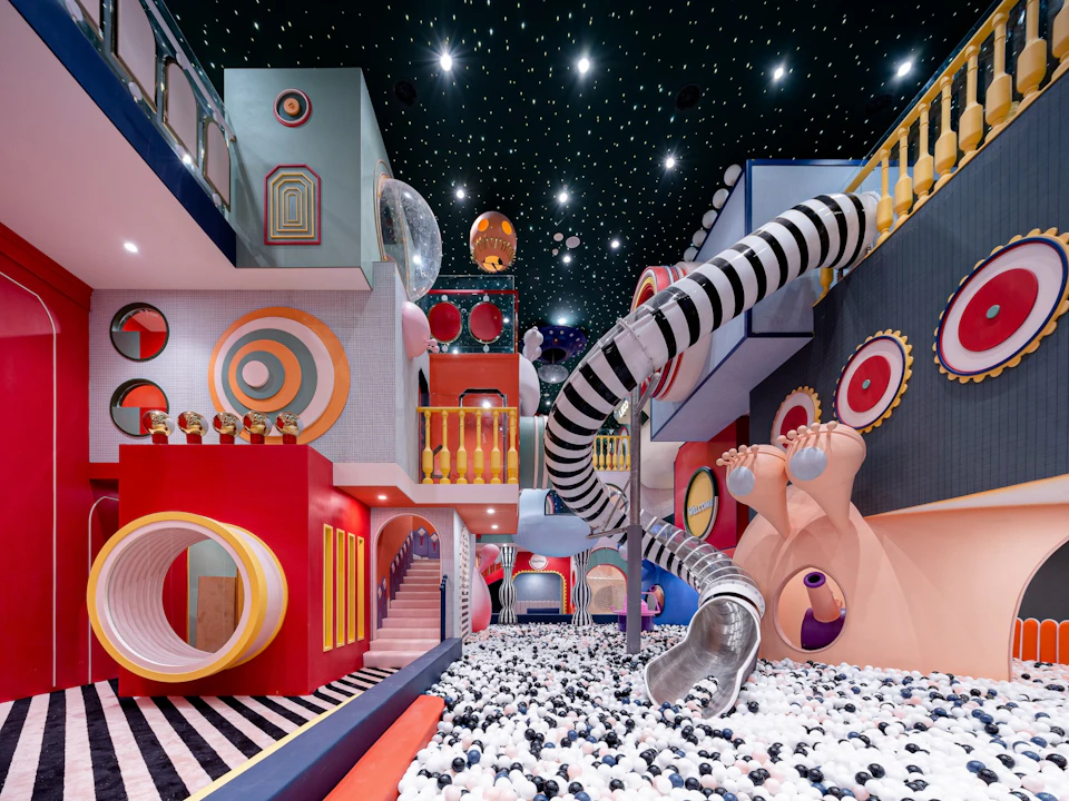 A colorful indoor playground with slides, ball pit, and artistic geometric decor under a ceiling with starry lights.