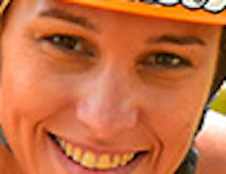 A person in red and yellow safety gear, wearing an orange helmet, smiles while strapped to a zip-line harness in a lush, green outdoor setting.