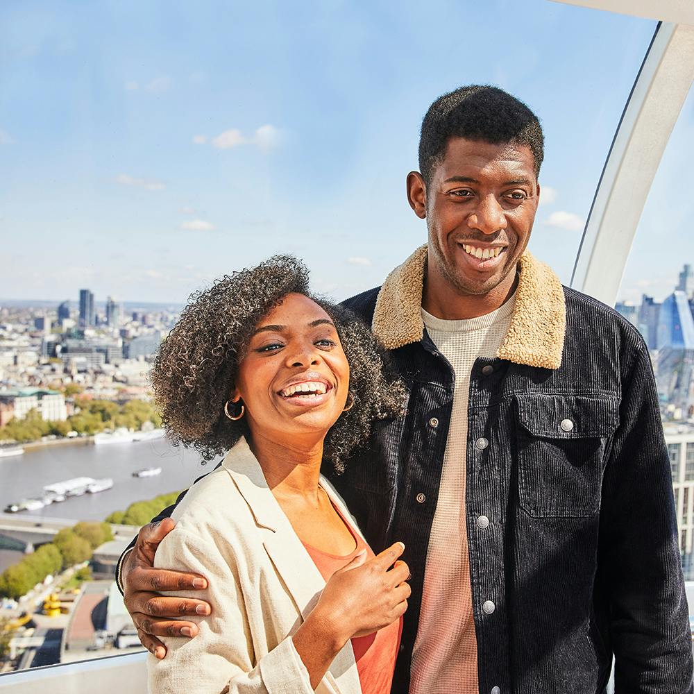 Usmívající se pár stojící uvnitř prosklené budovy s panoramatem města a řekou v pozadí.