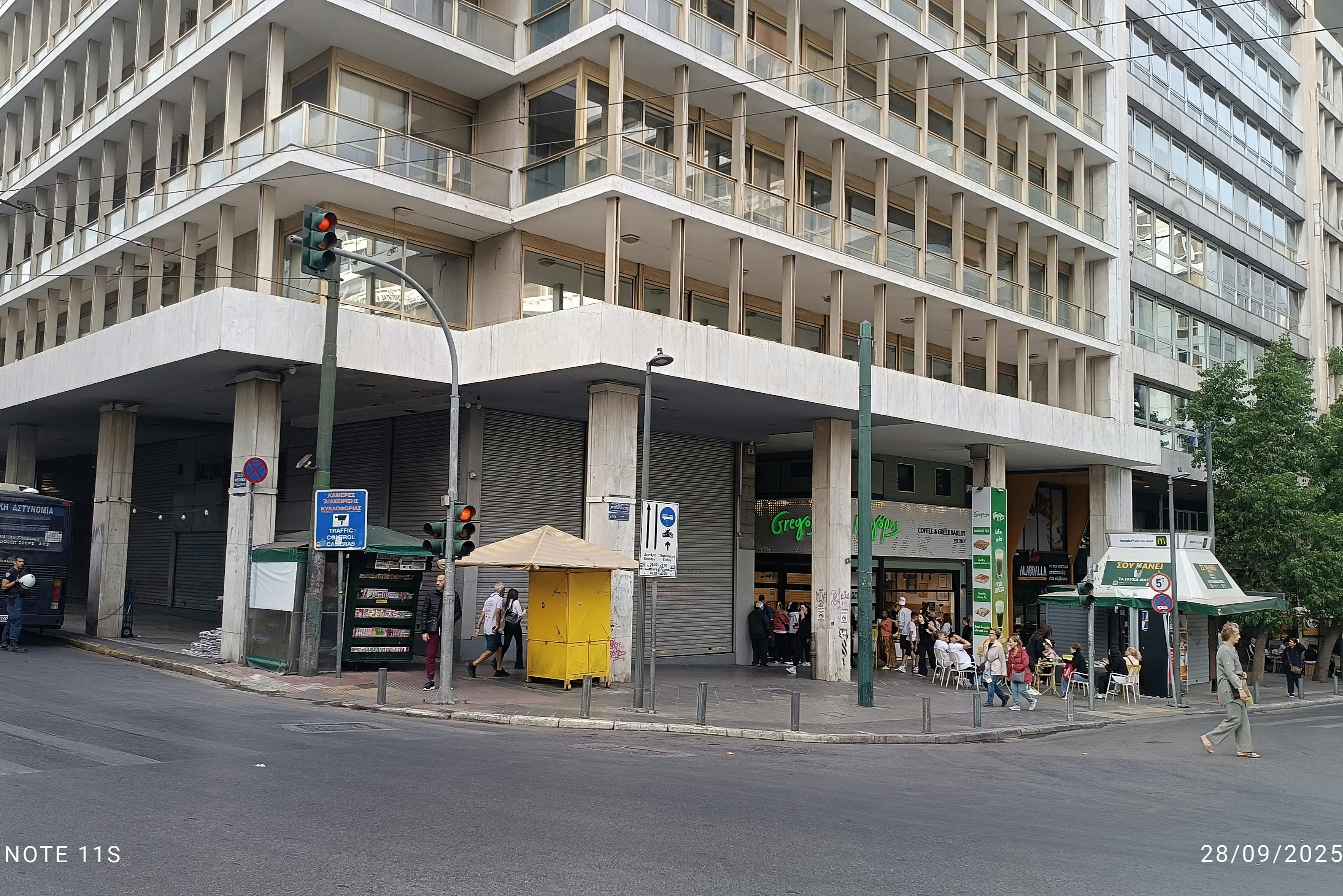 Οι άνθρωποι συγκεντρώνονται έξω από ένα κατάστημα στο ισόγειο ενός πολυώροφου κτιρίου, που βρίσκεται σε μια γωνία του δρόμου με ένα φανάρι.