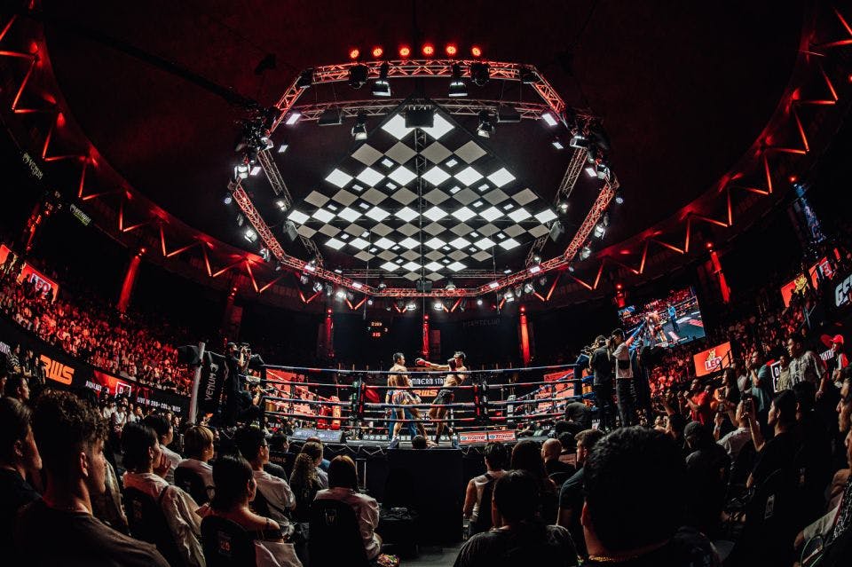 Un match de boxe dans une arène pleine à craquer, avec un éclairage dramatique et un plafond à carreaux. Les spectateurs et l'équipe de tournage encerclent le ring.
