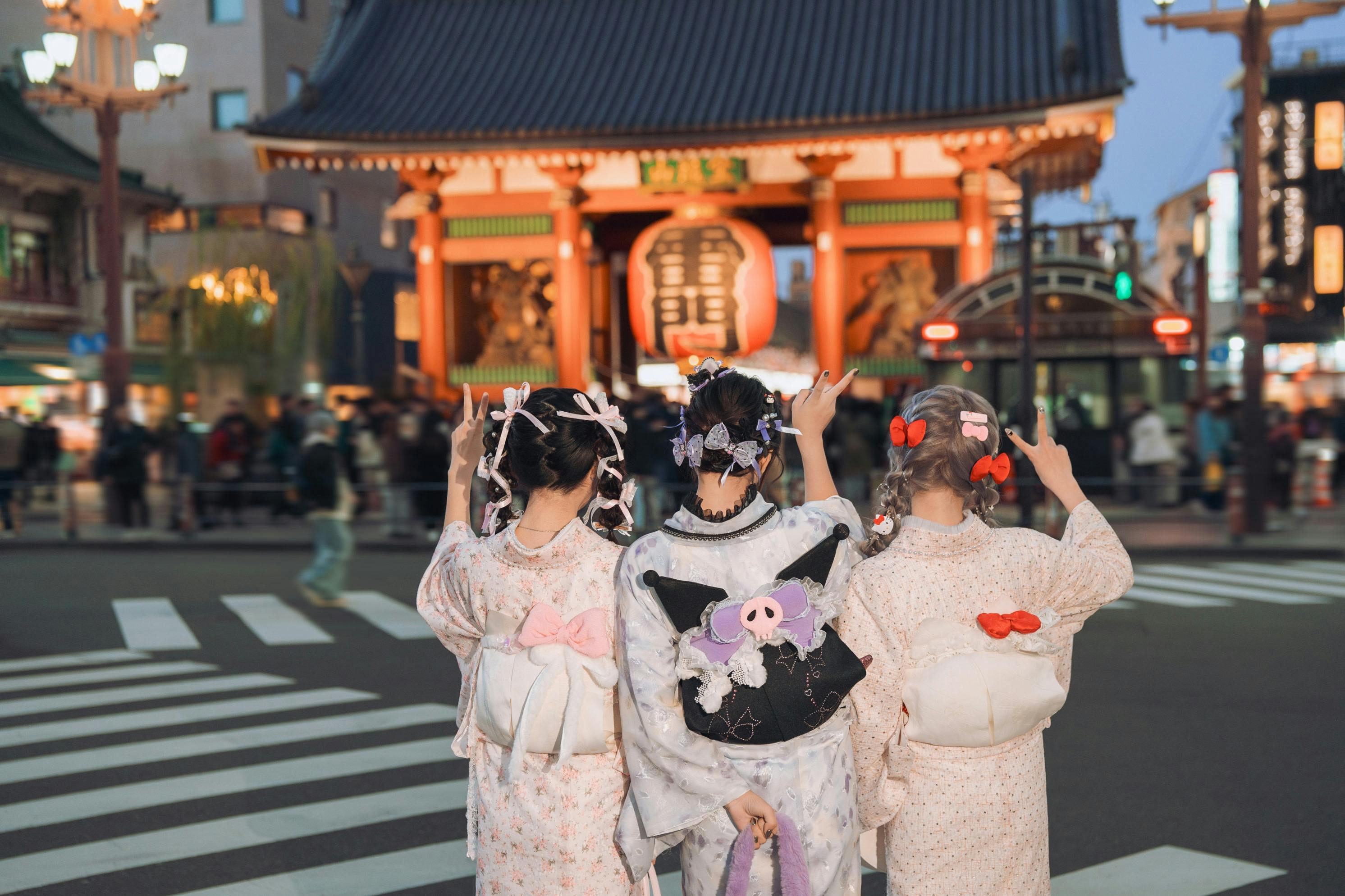 Three people in traditional attire stand with their backs facing the camera, gesturing, in front of a large red temple gate.