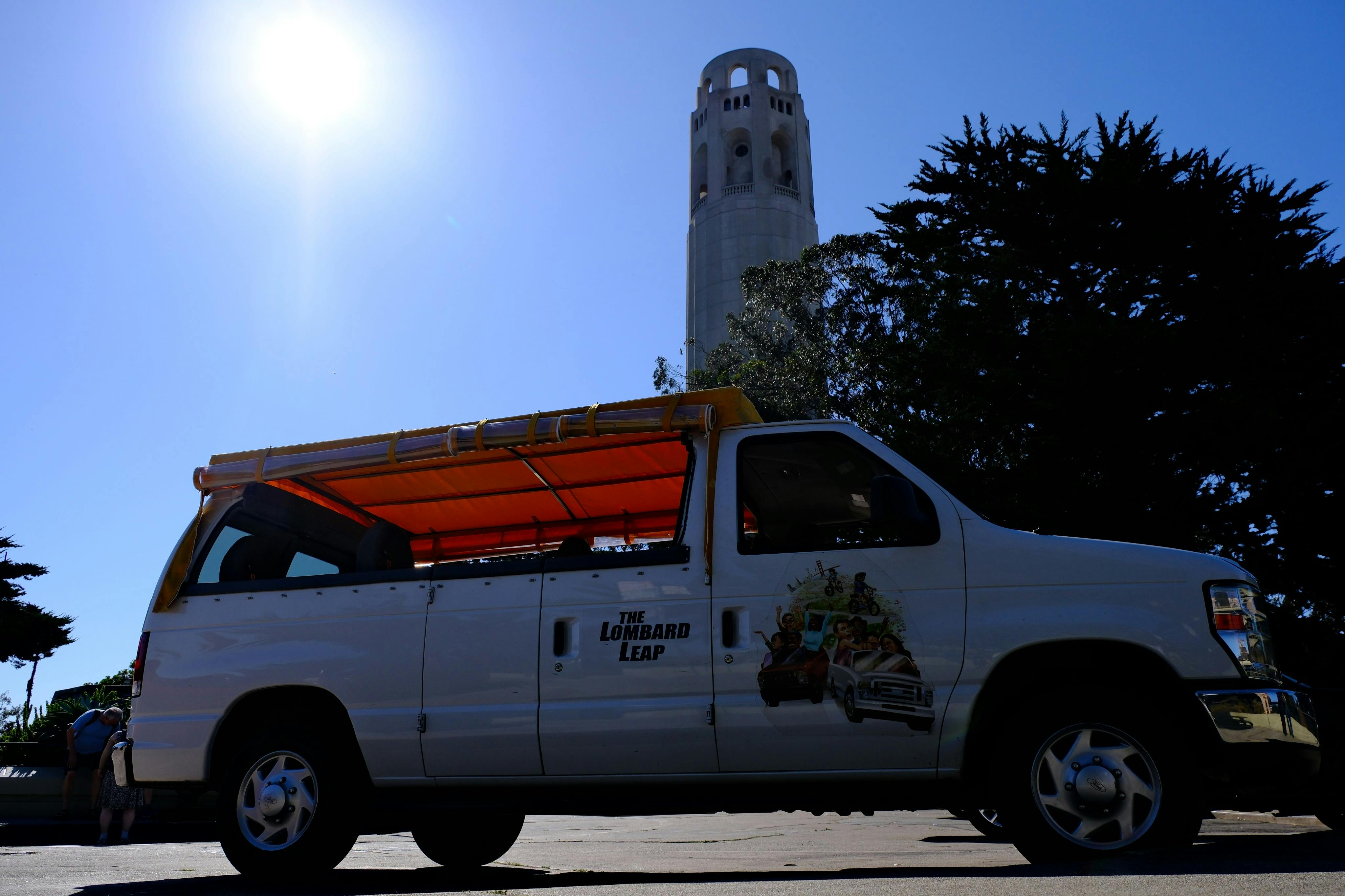 A white van with "The Lombard Leap" branding is parked near trees, with a tall tower and the sun visible in the background.