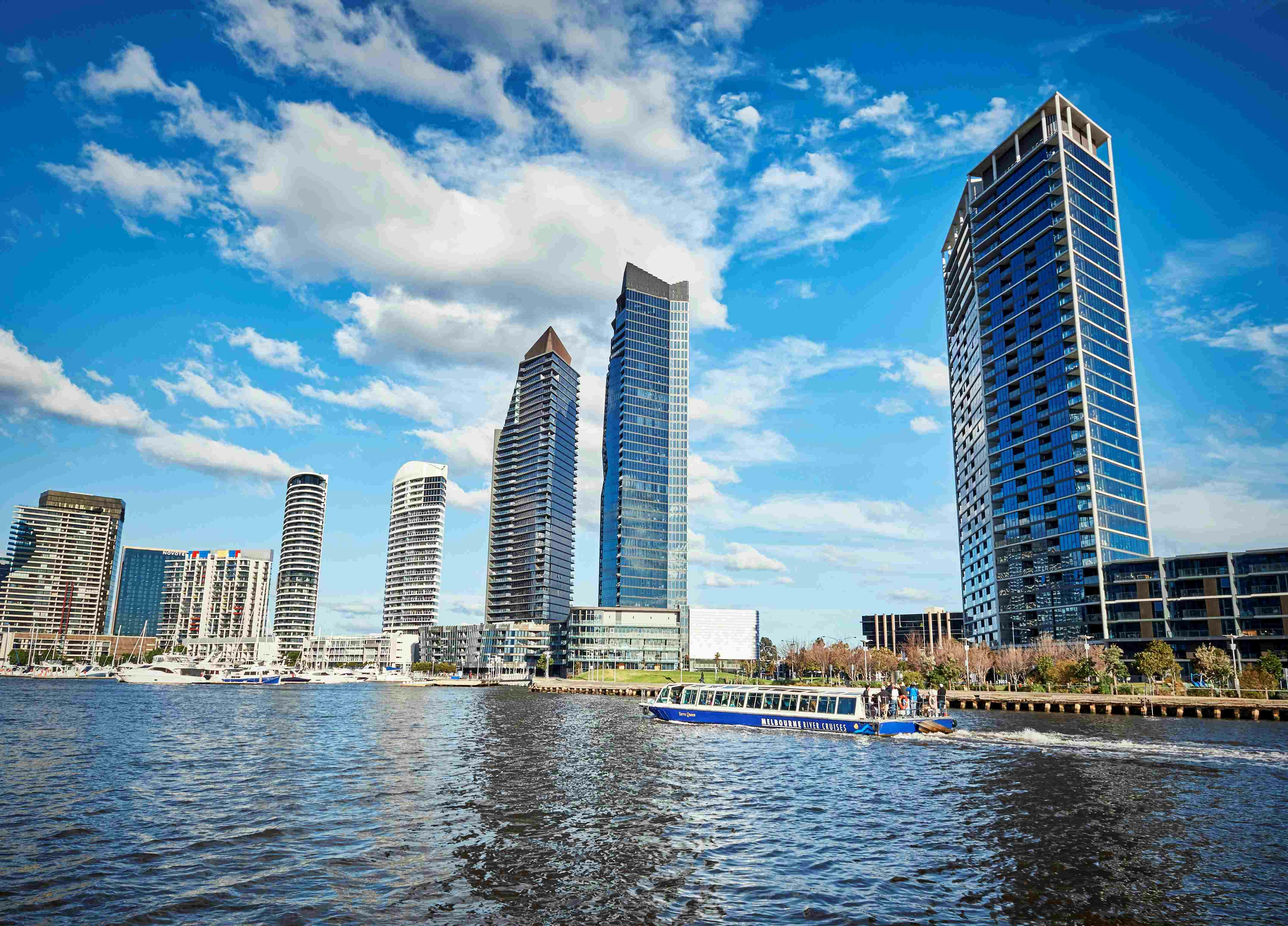 Αστικός ορίζοντας με ψηλούς, μοντέρνους ουρανοξύστες που αντανακλούν σε μια προκυμαία και ένα μπλε τουριστικό σκάφος που πλοηγείται στο νερό.