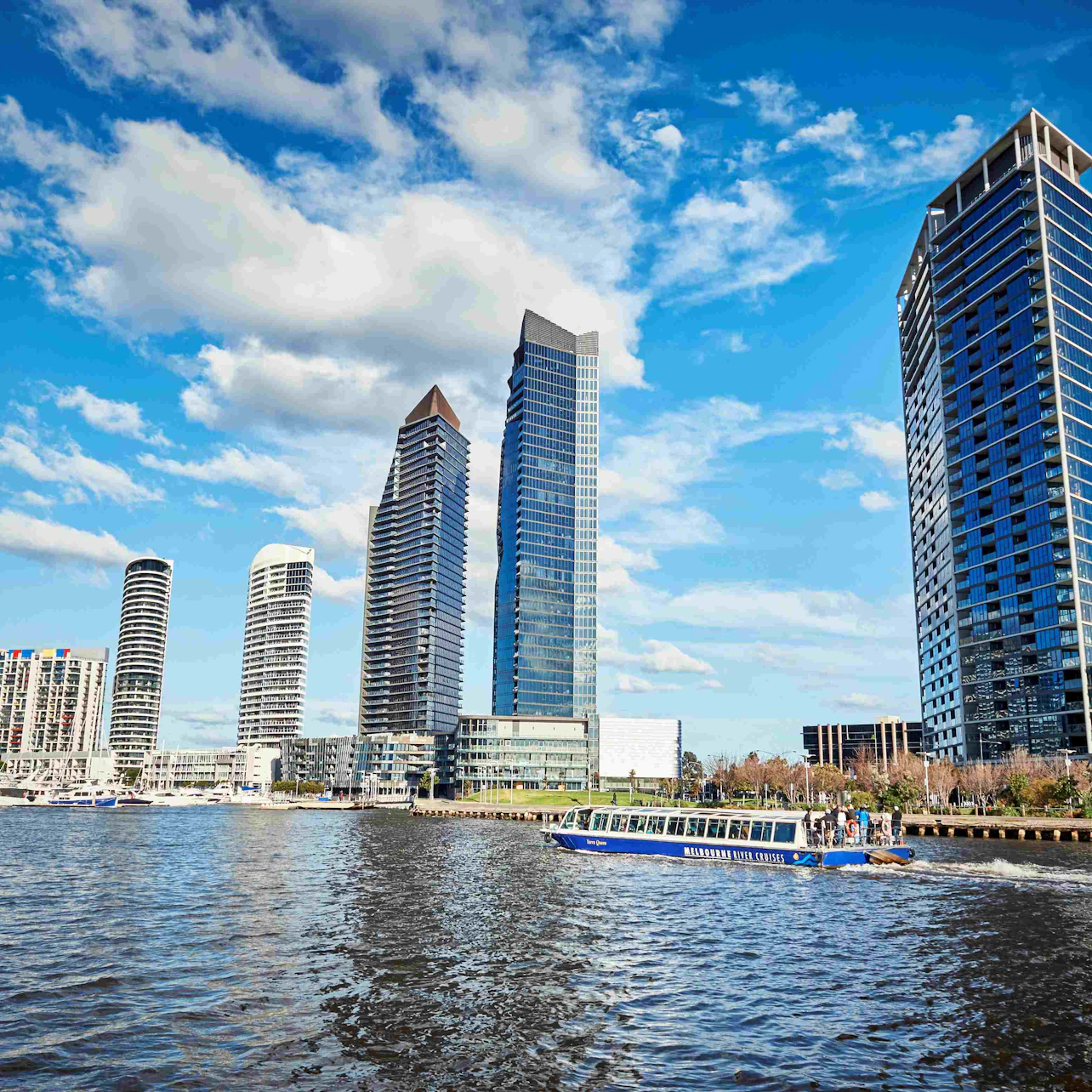 Urban skyline with tall, modern skyscrapers reflecting on a waterfront, and a blue tour boat navigating the water.