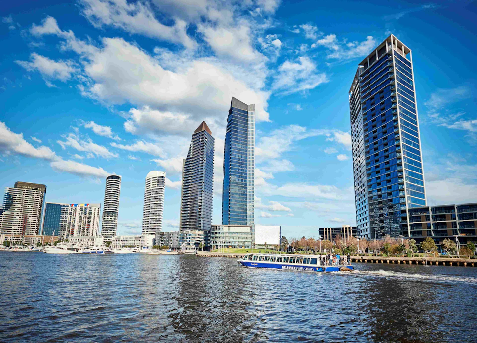 Urban skyline with tall, modern skyscrapers reflecting on a waterfront, and a blue tour boat navigating the water.