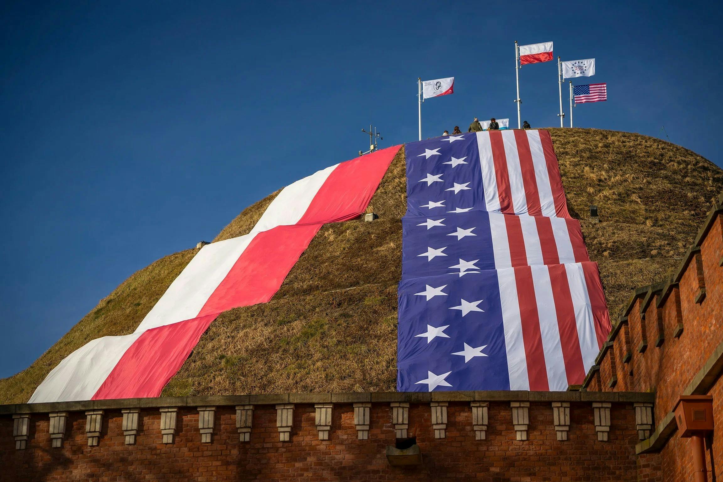 A hill adorned with large flags resembling the Polish and American flags, with smaller flags on poles at the top under a blue sky.