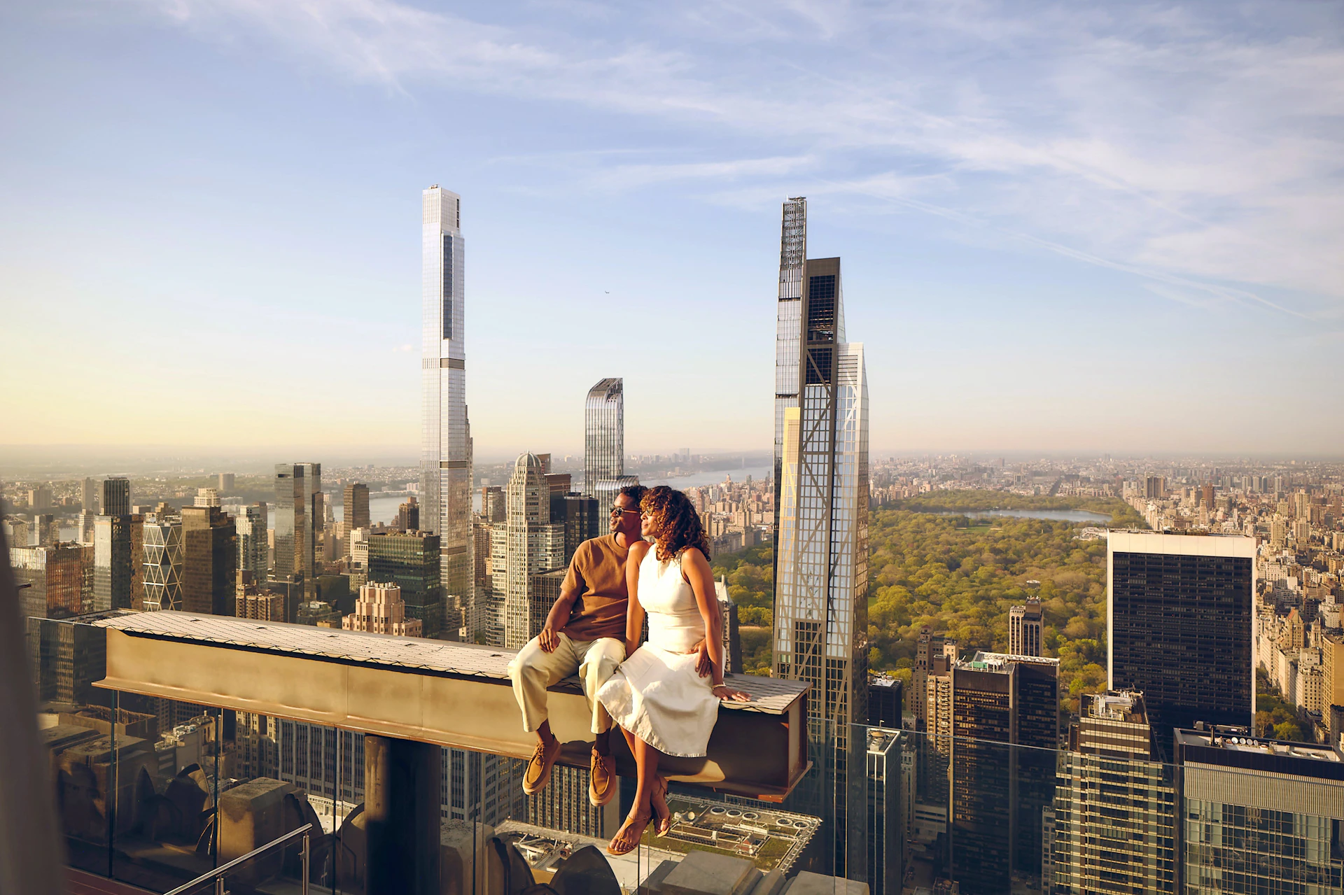 A couple sits on a beam atop a skyscraper, overlooking a cityscape with tall buildings and a distant park under a blue sky.