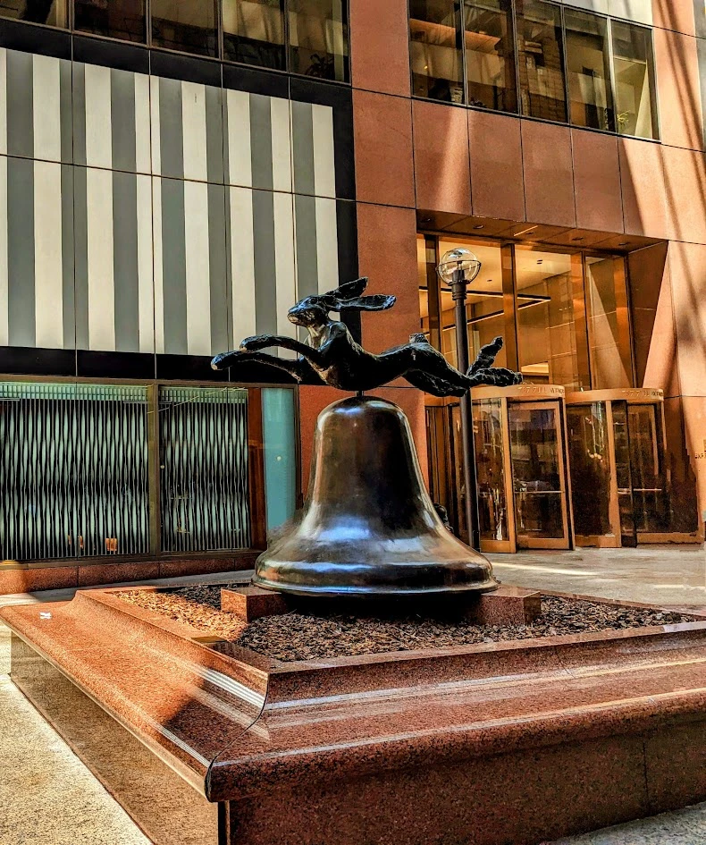 A bronze sculpture of a hare leaping over a bell, placed in a plaza with a modern building facade in the background.