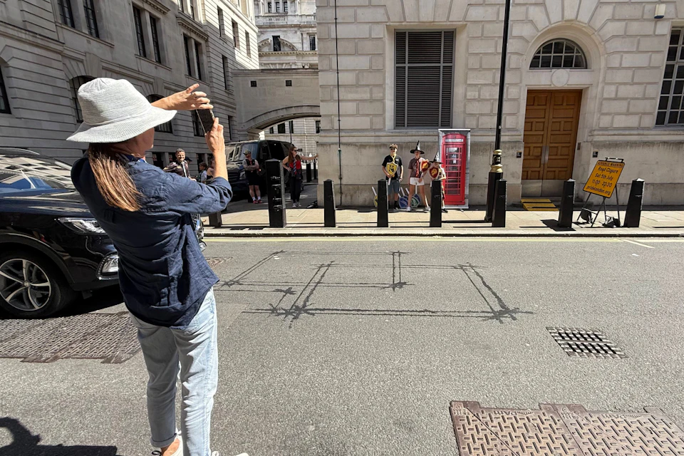 A person in a hat taking a photo of a group posing near a red phone booth on a sunny city street.