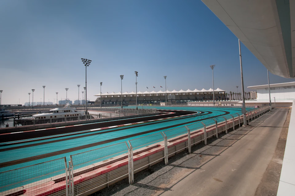 A curved racing track with turquoise and black lanes, under a clear blue sky, with a grandstand and floodlights in the background.