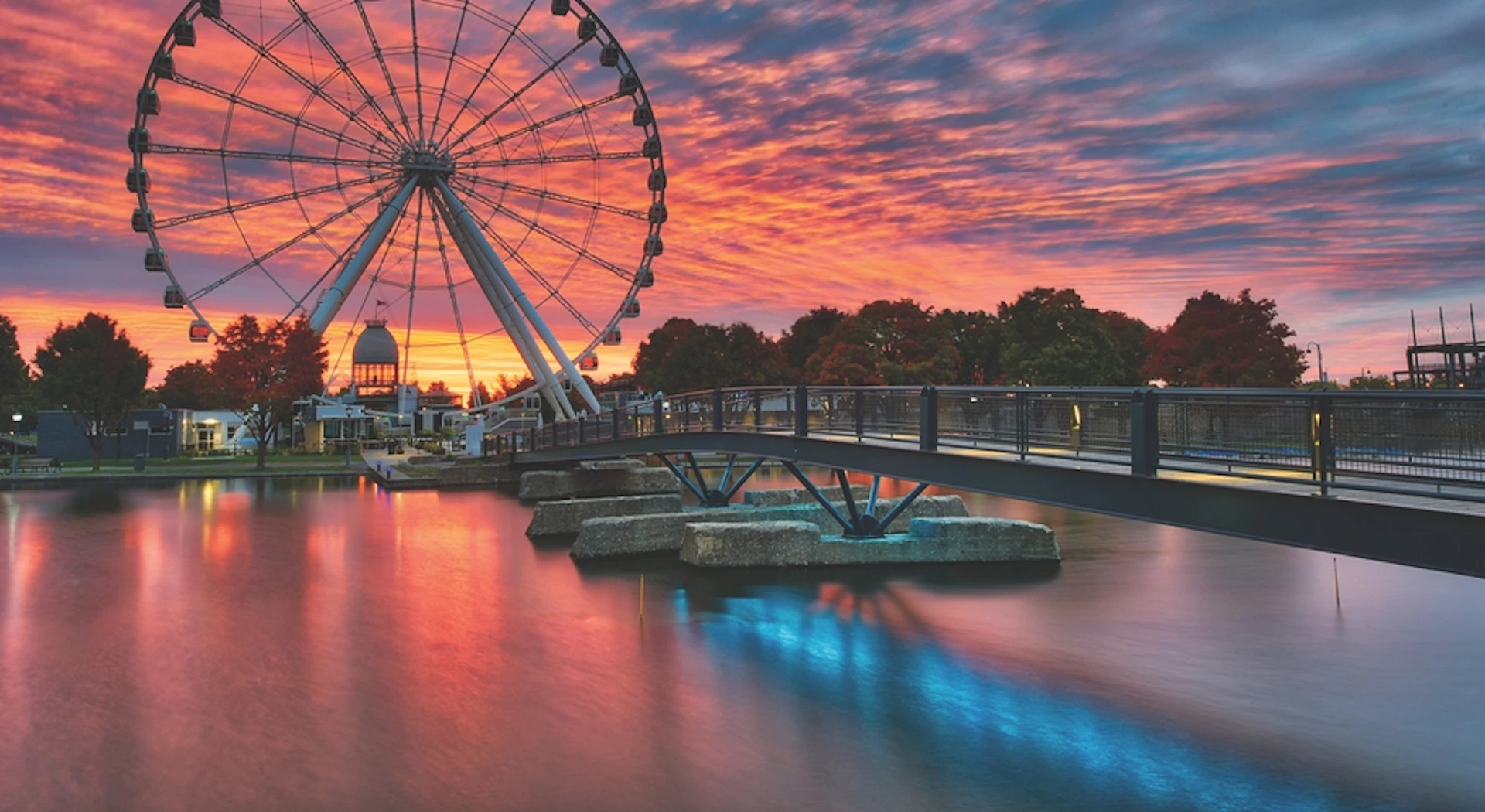 Ferris wheel by a bridge over water, with a vibrant sunset sky and trees in the background.