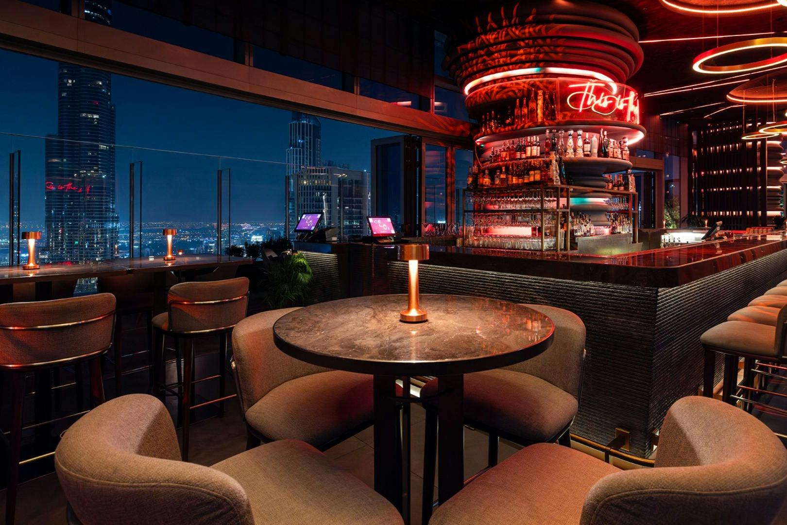 Sophisticated rooftop bar with city skyline view, cozy seating around a round table, mood lighting, and a well-stocked modern bar.