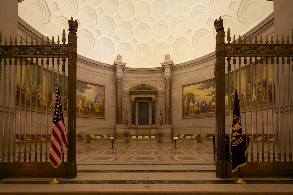 A grand hall with a domed ceiling, large paintings, American flags, and ornate gates.