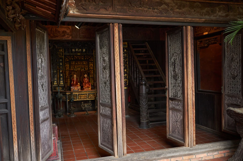 An interior view of a traditional wooden building with ornate doors, tiled floor, a staircase, and a decorative altar.