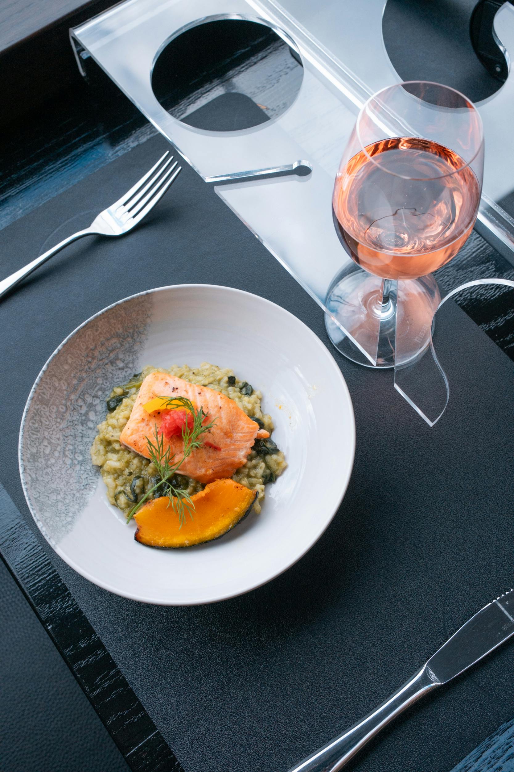 Een bord zalm op risotto, gegarneerd met kruiden en pompoen, met een vork en een glas rosé ernaast.