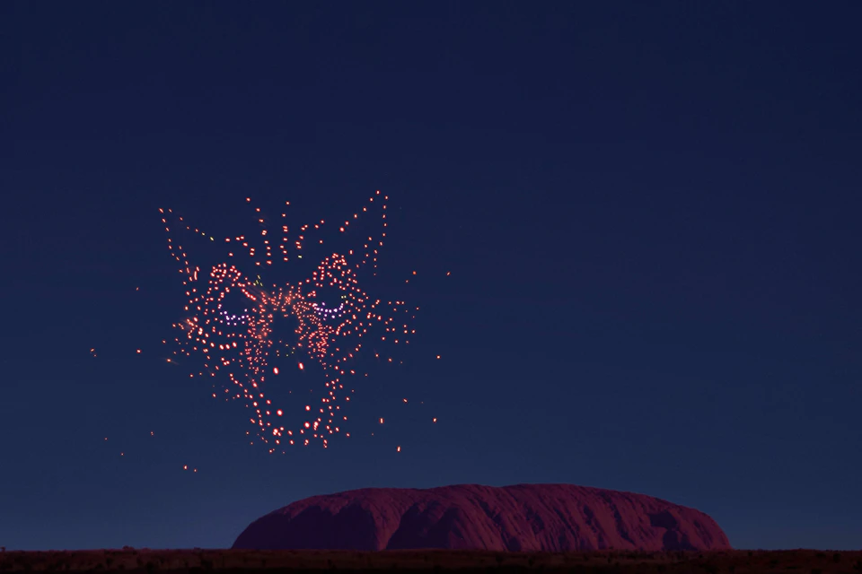 A night sky shows drones forming a glowing wolf face over a large rock formation, resembling Uluru, silhouetted by moonlight.