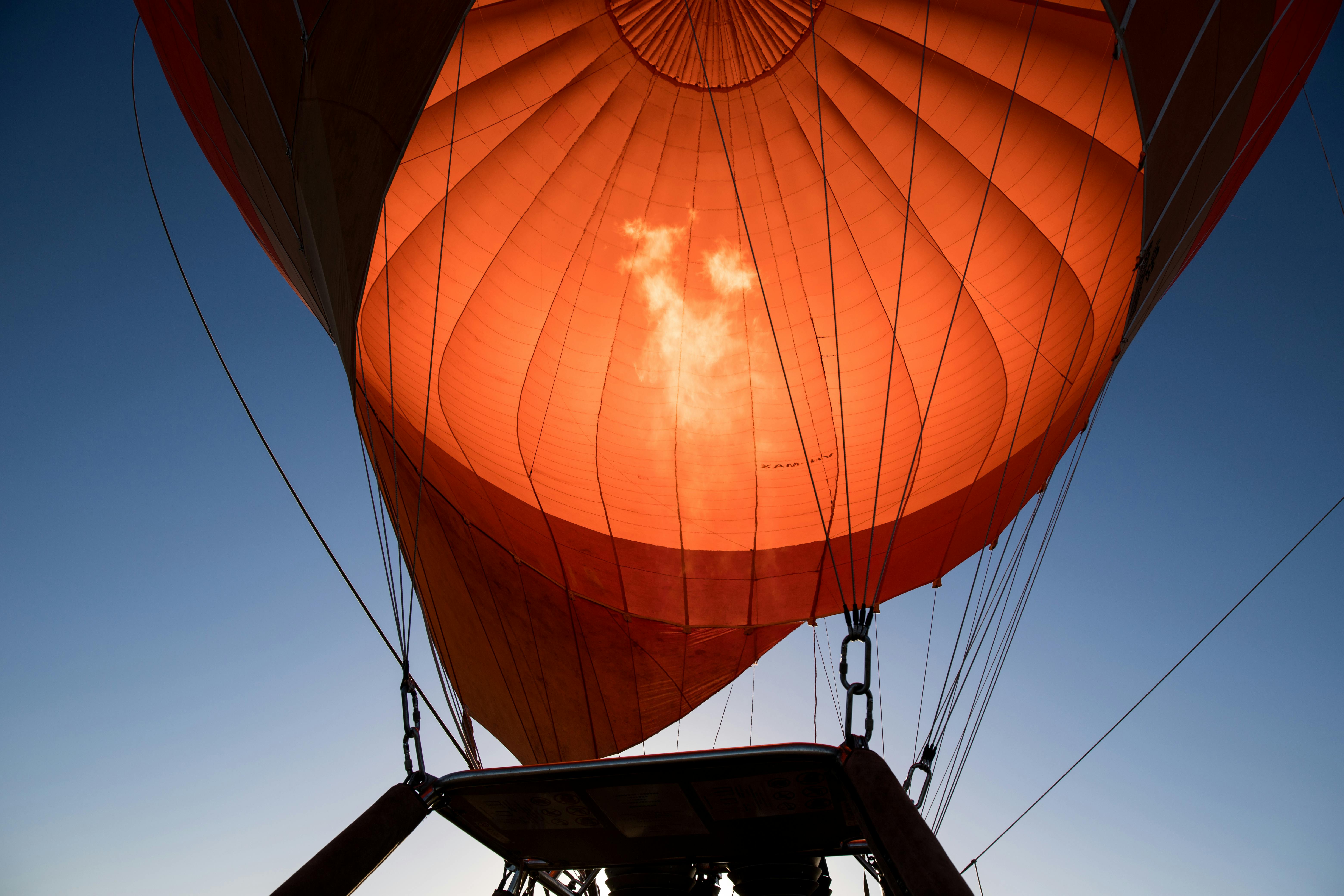 Pohled zevnitř koše horkovzdušného balónu, pohled na osvětlený oranžový baldachýn proti modré obloze.
