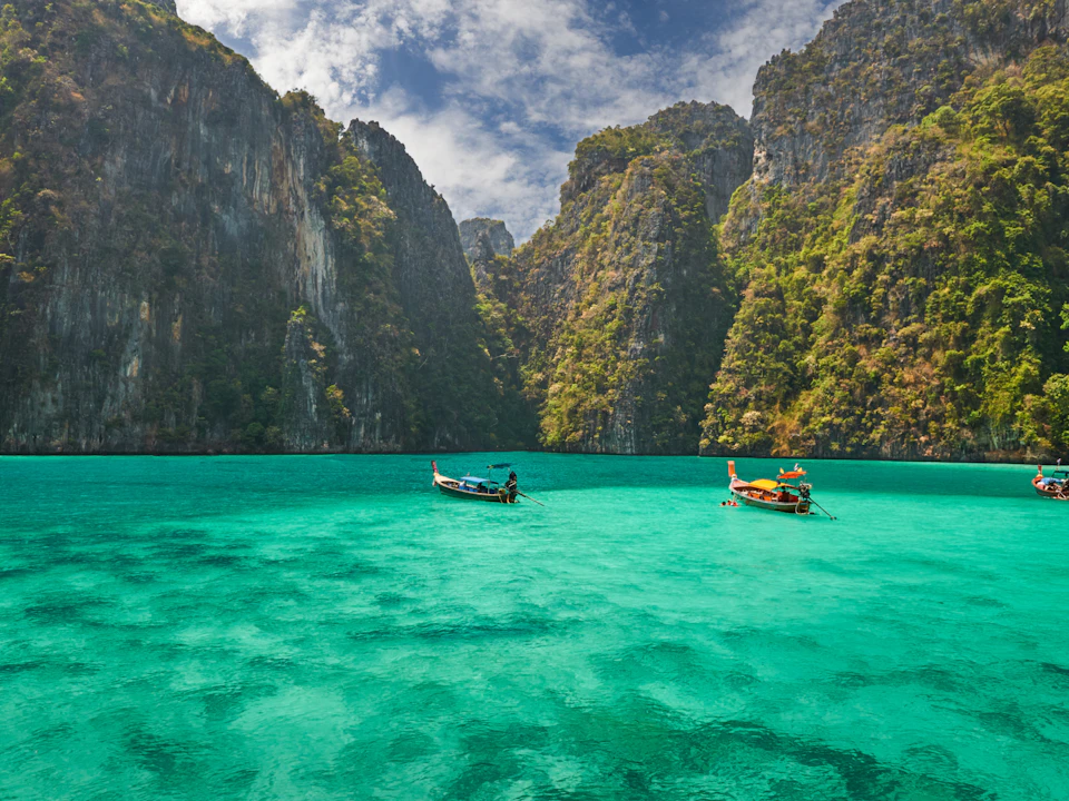 Two small boats floating on clear turquoise water, surrounded by tall, lush green cliffs under a partially cloudy sky.