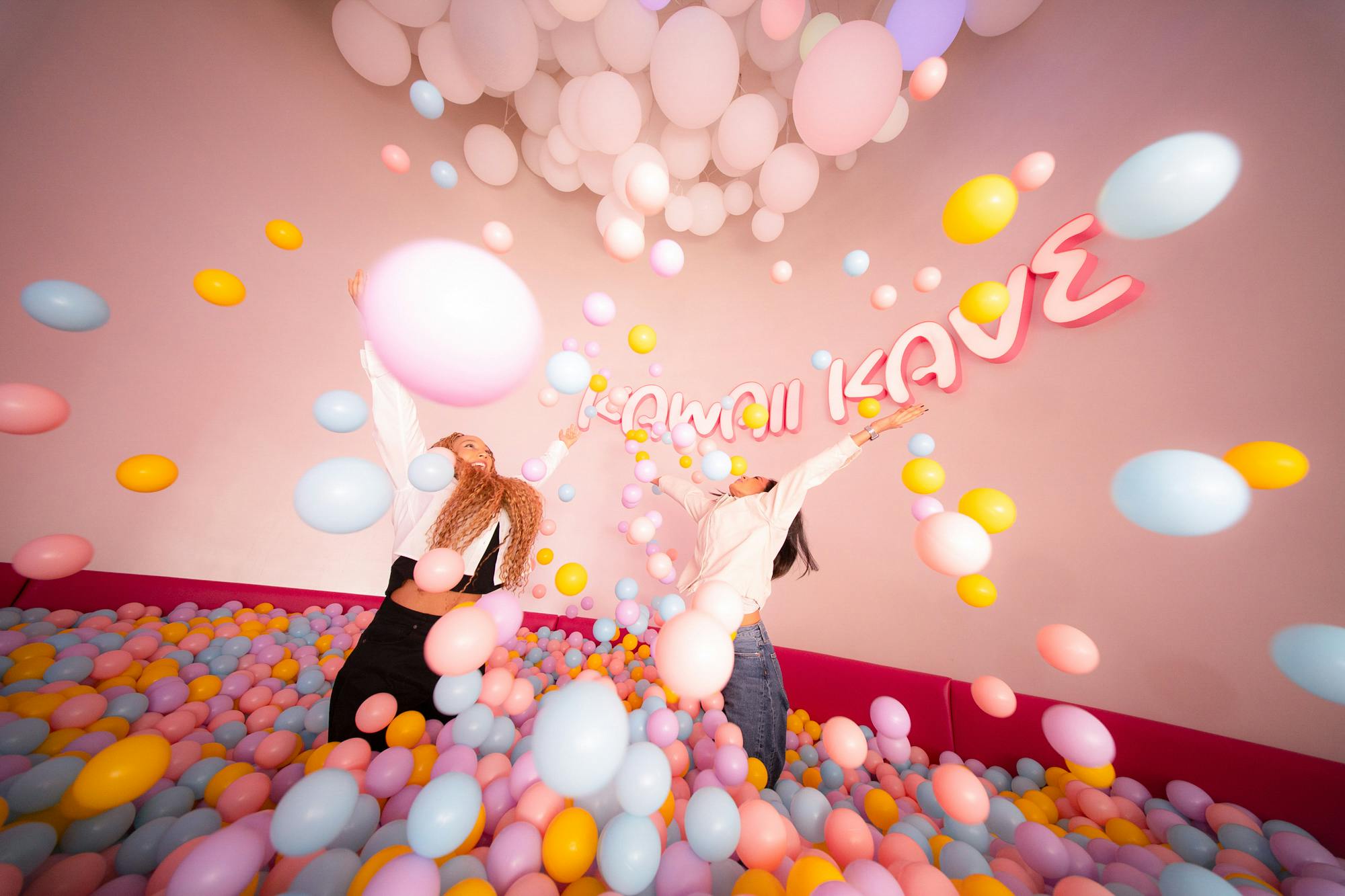 하얀 풍선 아래 벽에 "KAWAII Kave" 간판이 있는 화려한 공 구덩이에 두 사람이 즐겁게 파스텔 풍선을 던집니다.