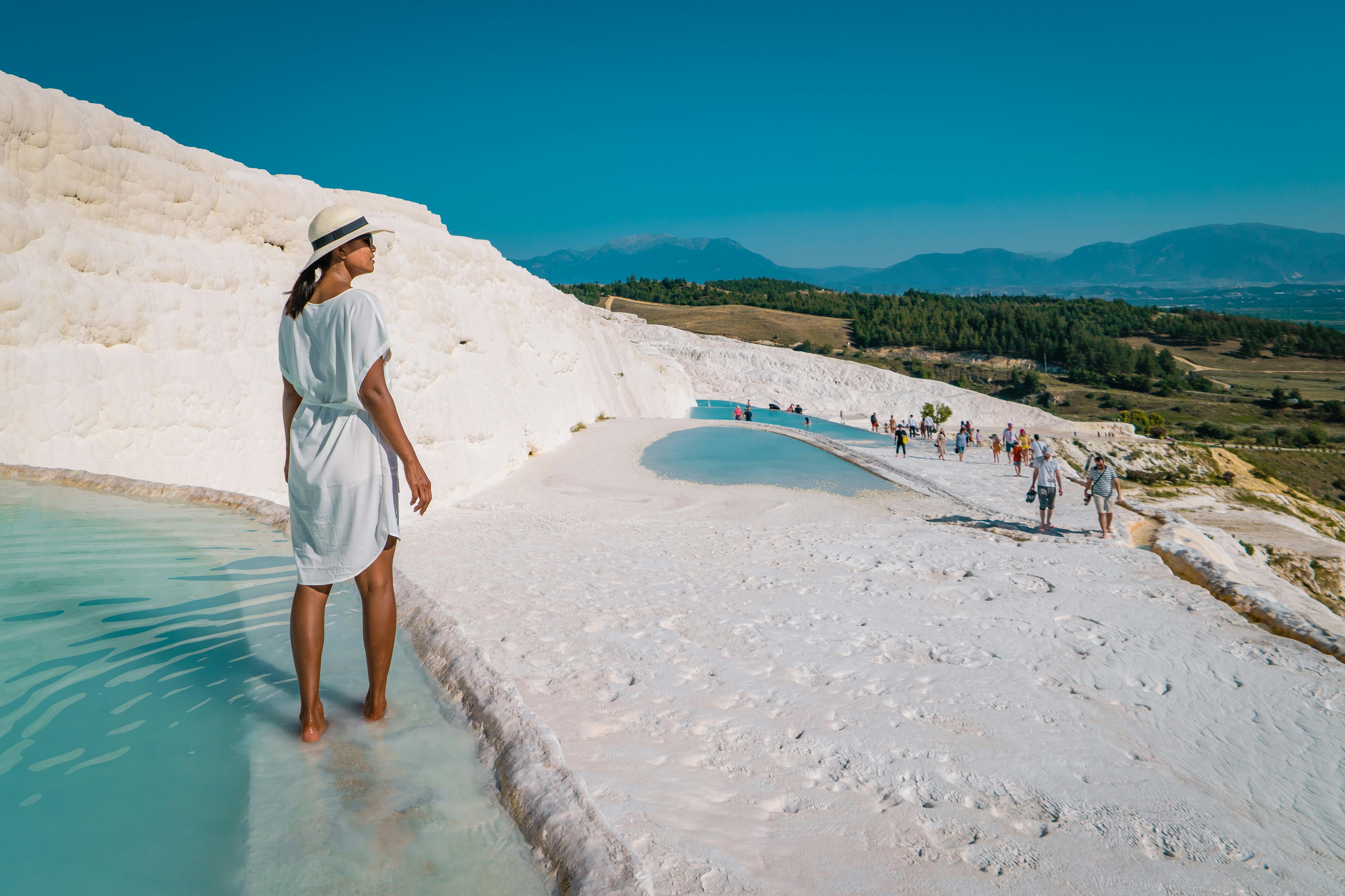 A woman in a white dress and hat walks through shallow turquoise water surrounded by white travertine terraces and hills.