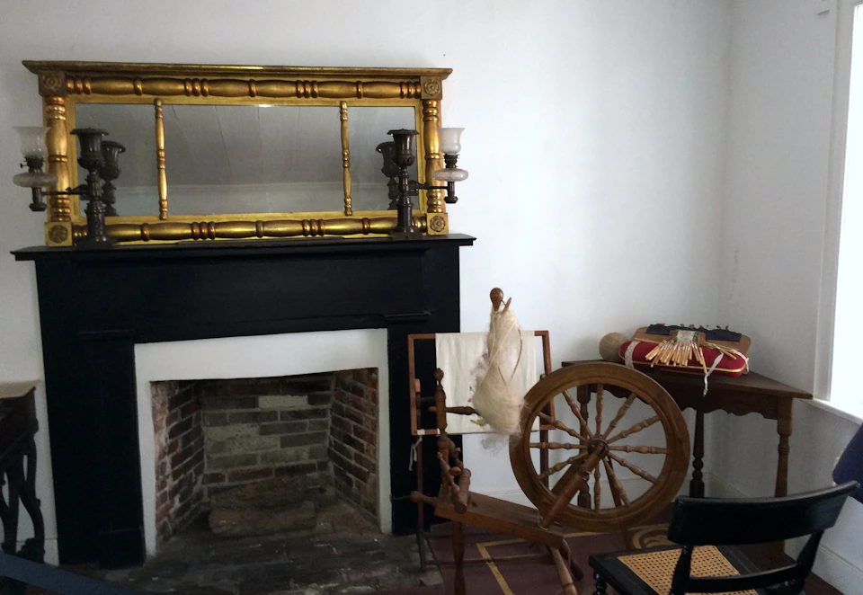 Room with a black fireplace, a spinning wheel, and a large golden mirror above the mantle. Table with yarn in the background.