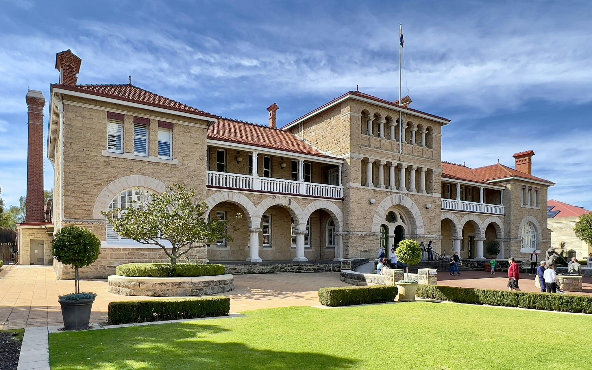 A beige brick building with arched windows and balconies, surrounded by a garden with people walking around.