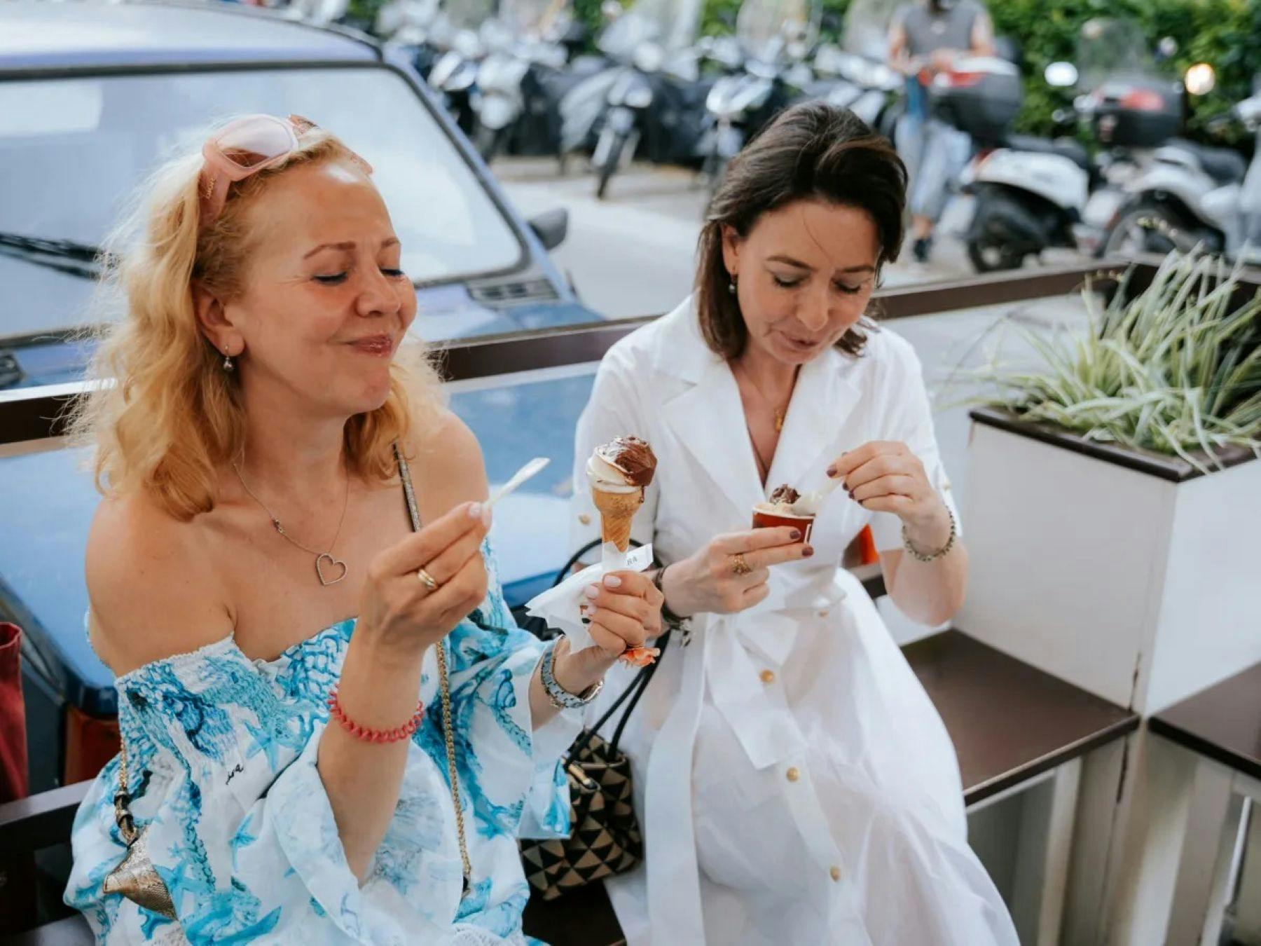 Due donne sedute all'aperto che si godono un gelato, con degli scooter parcheggiati visibili sullo sfondo.