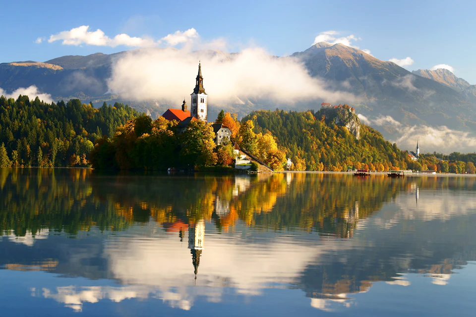 A church with a tall steeple on a lush, forested island, reflected in a calm lake, with mountains and a cloudy sky in the background.