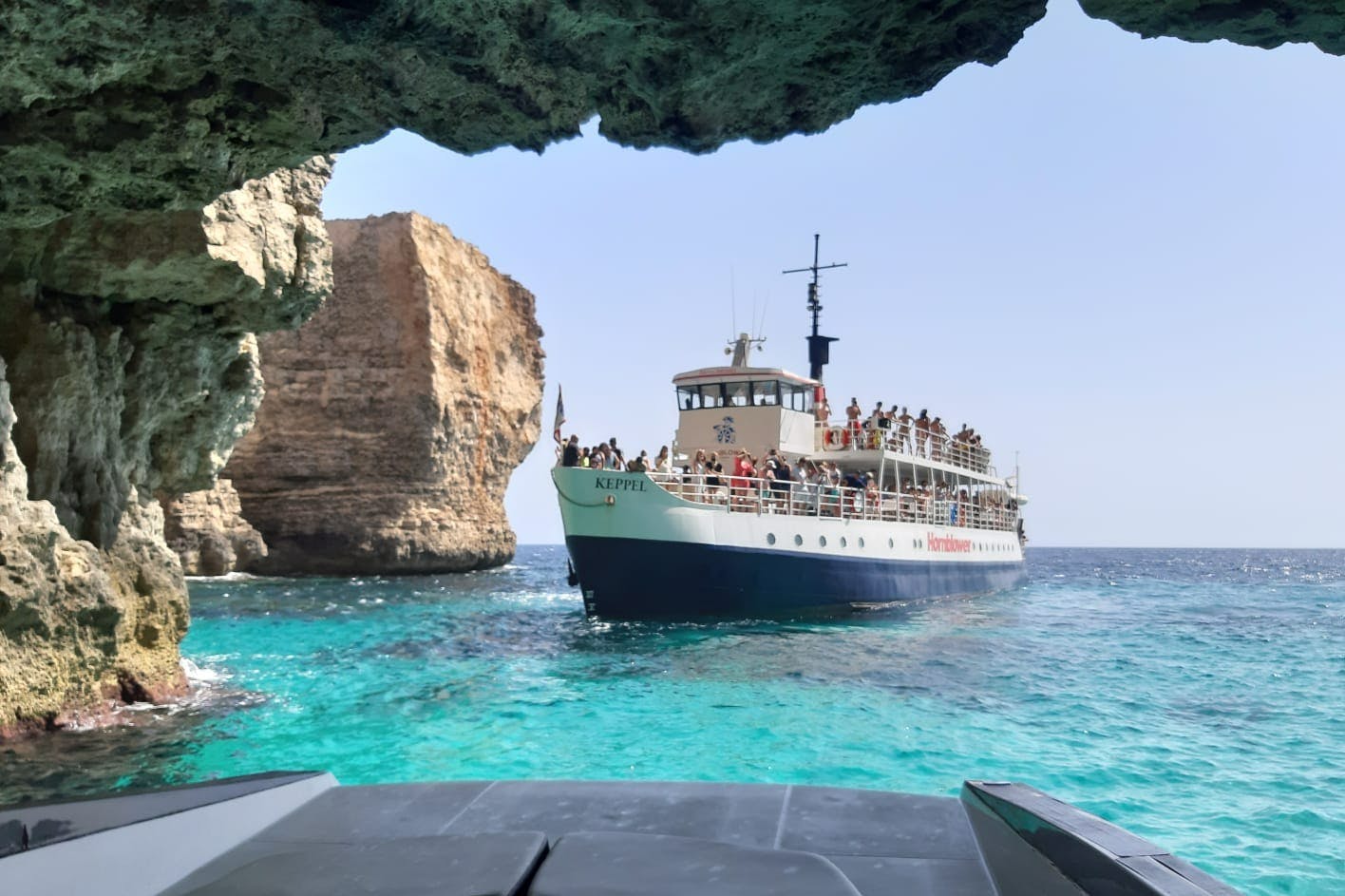 Ένα σκάφος που ονομάζεται «Keppel» με επιβάτες επί του σκάφους πλέει σε καθαρά γαλαζοπράσινα νερά κοντά σε ένα σχηματισμό βράχων κάτω από την άκρη μιας σπηλιάς.