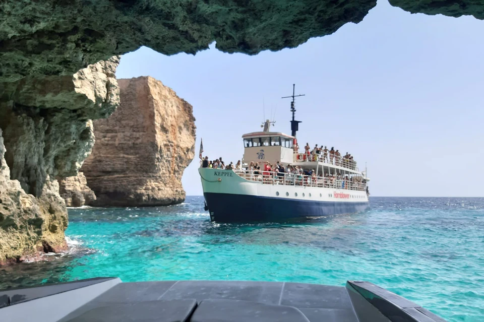 A boat named 'Keppel' with passengers on board is sailing in clear turquoise water near a rock formation under a cave's edge.