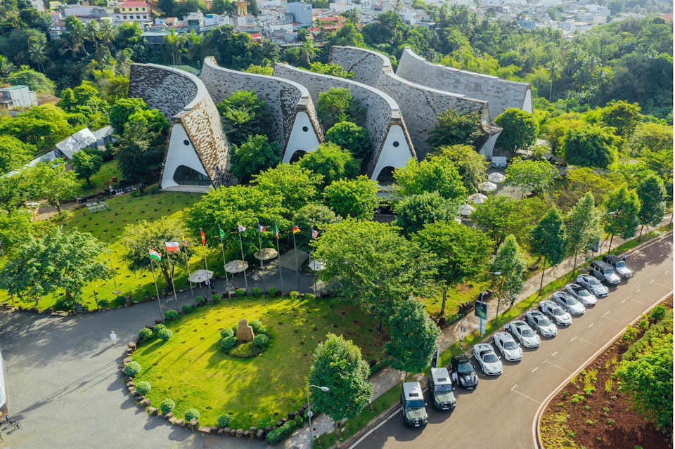 Aerial view of a modern building with curved roofs surrounded by greenery, flags, and parked cars in a city setting.