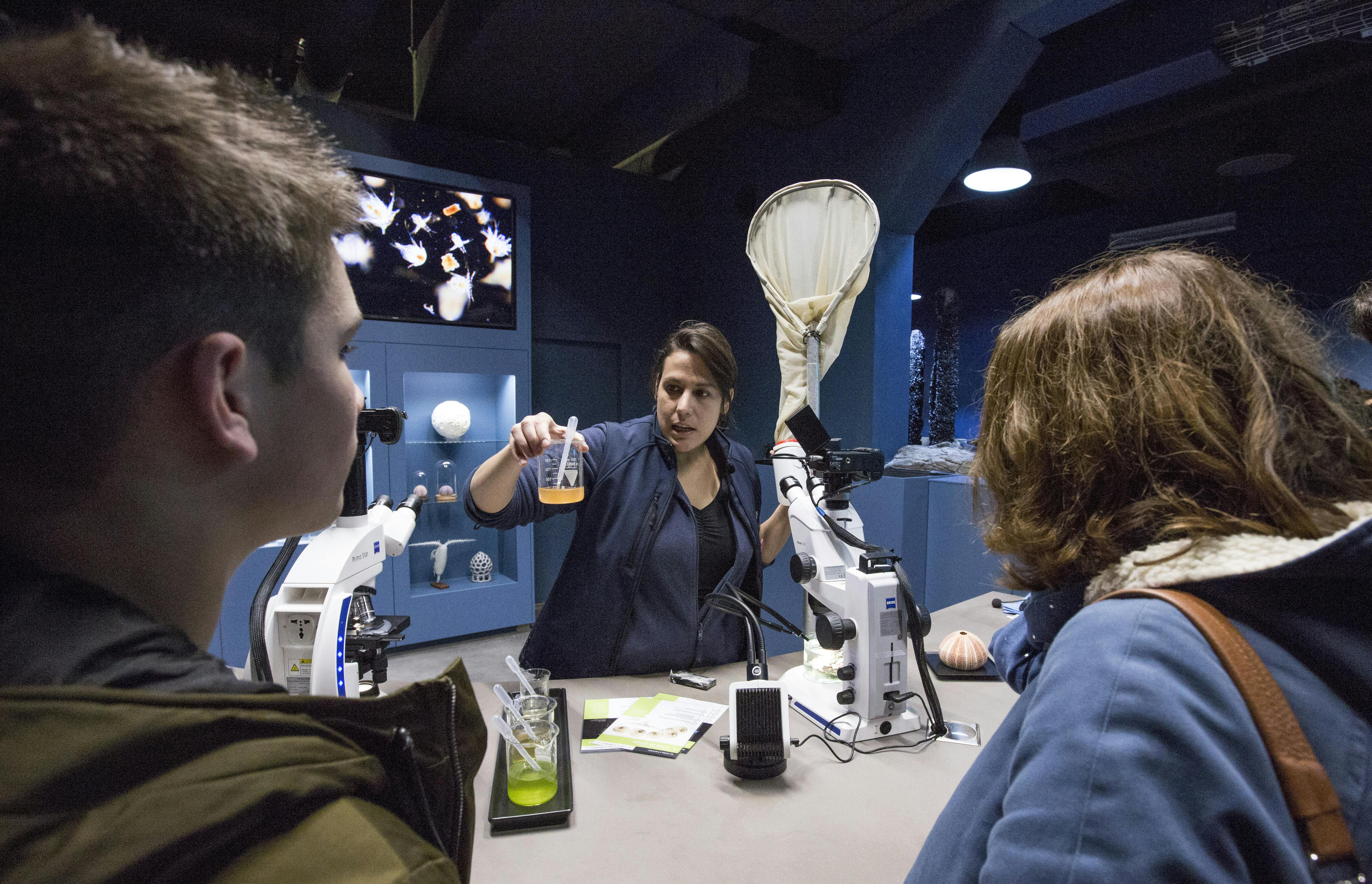 A woman demonstrates lab equipment to two people in a dimly lit room with microscopes, scientific tools, and screens in the background.