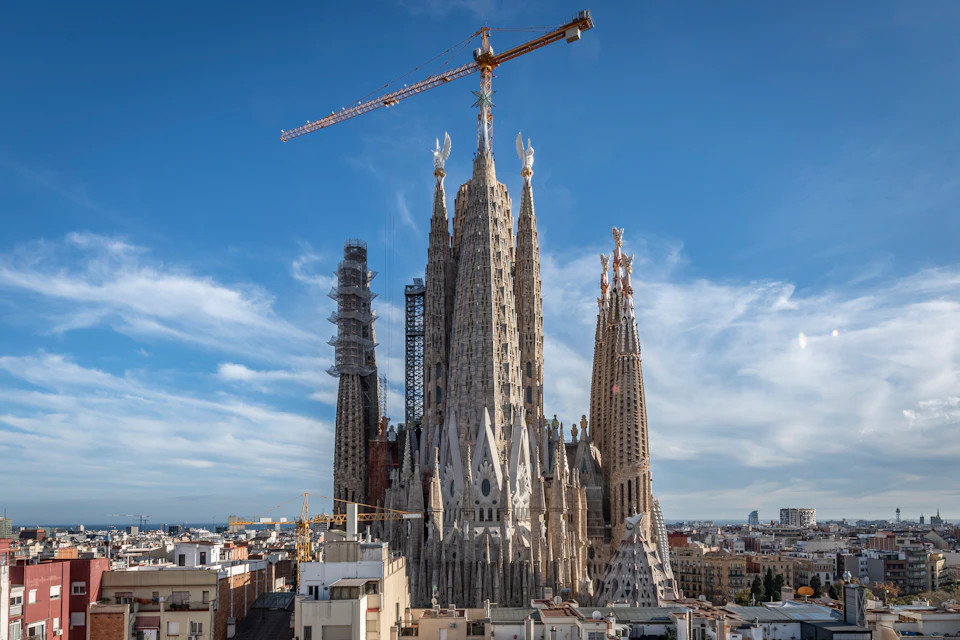 A large cathedral with multiple spires under construction, featuring cranes, against a cityscape and blue sky backdrop.