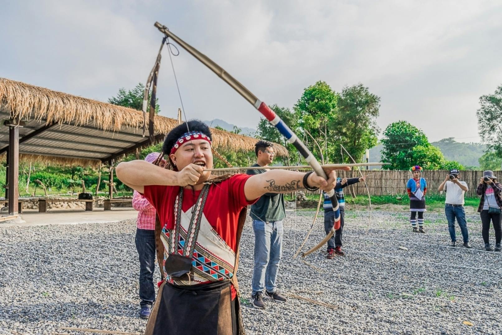 A person outdoors drawing a bow and arrow, wearing traditional attire, with others and greenery in the background.