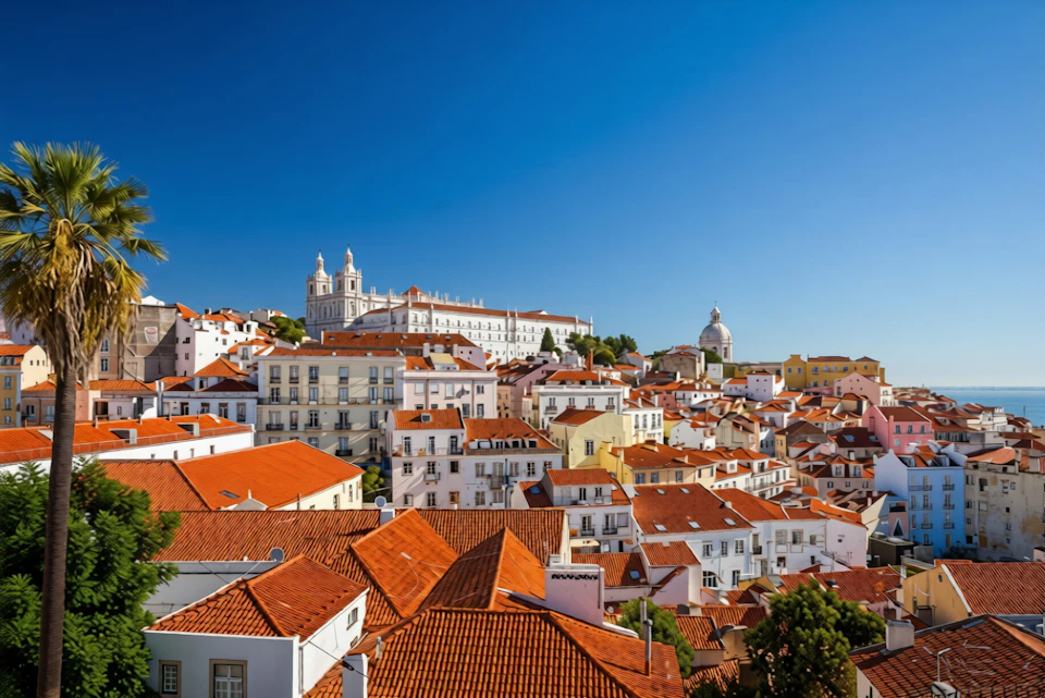 A hilly cityscape with numerous buildings featuring red-tiled roofs, a white church, and a clear blue sky.