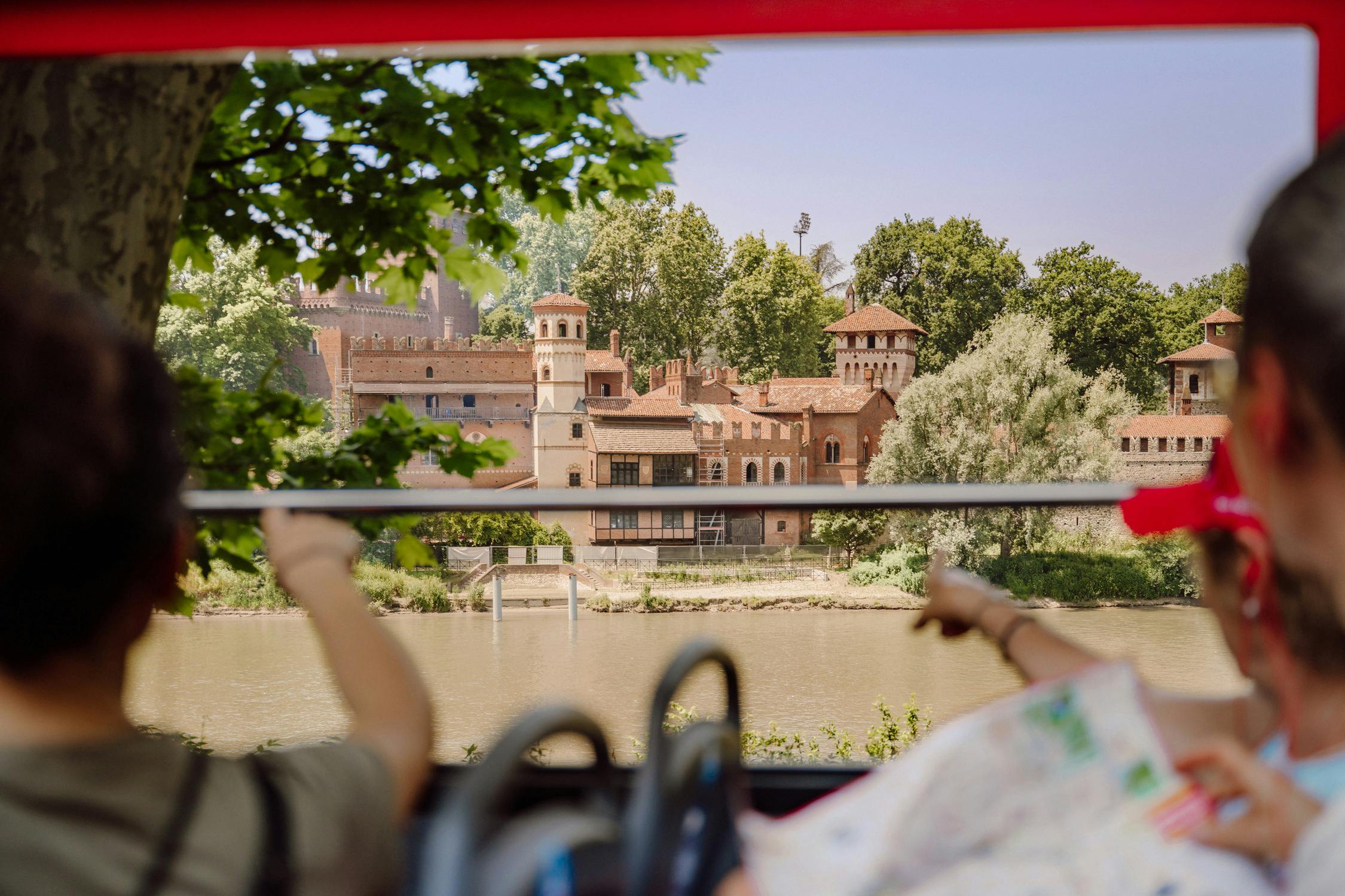 A scenic view of a riverside castle-like building with trees behind, viewed from a moving vehicle with passengers pointing.
