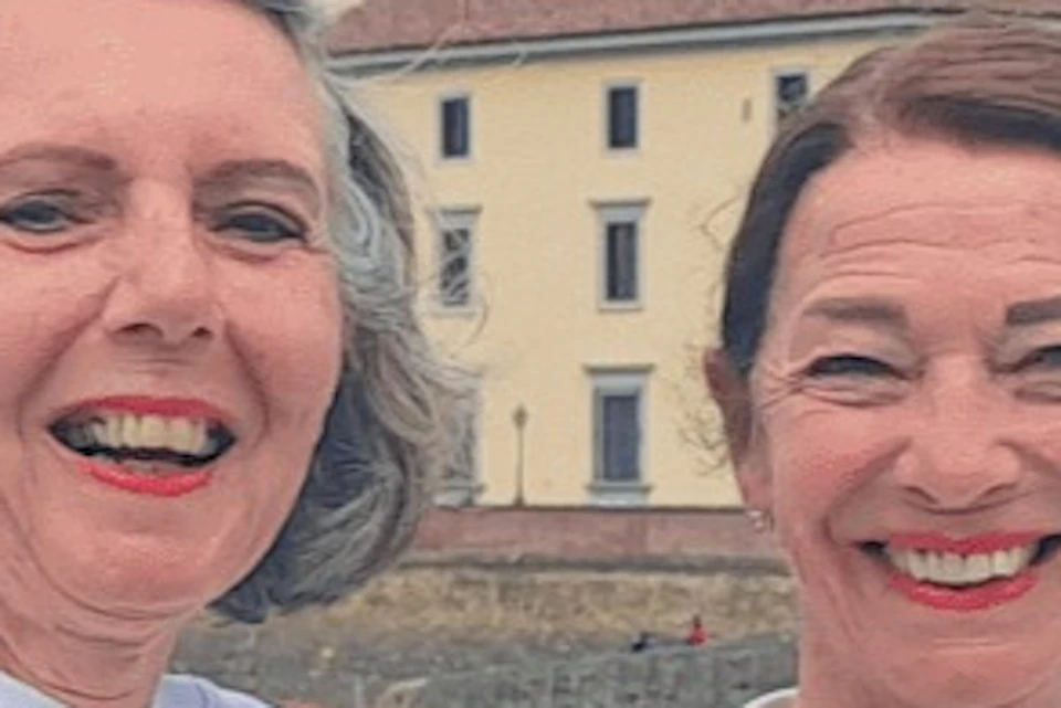 Two women standing on a bridge, smiling and giving thumbs-up, with a riverside town and construction cranes in the background.