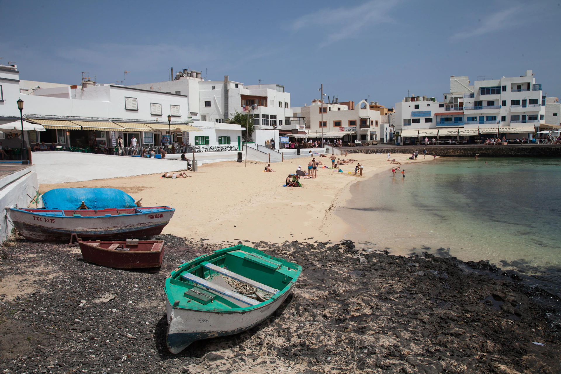 Small boats on rocky shore, people relaxing on sandy beach, and white buildings in the background under a blue sky.
