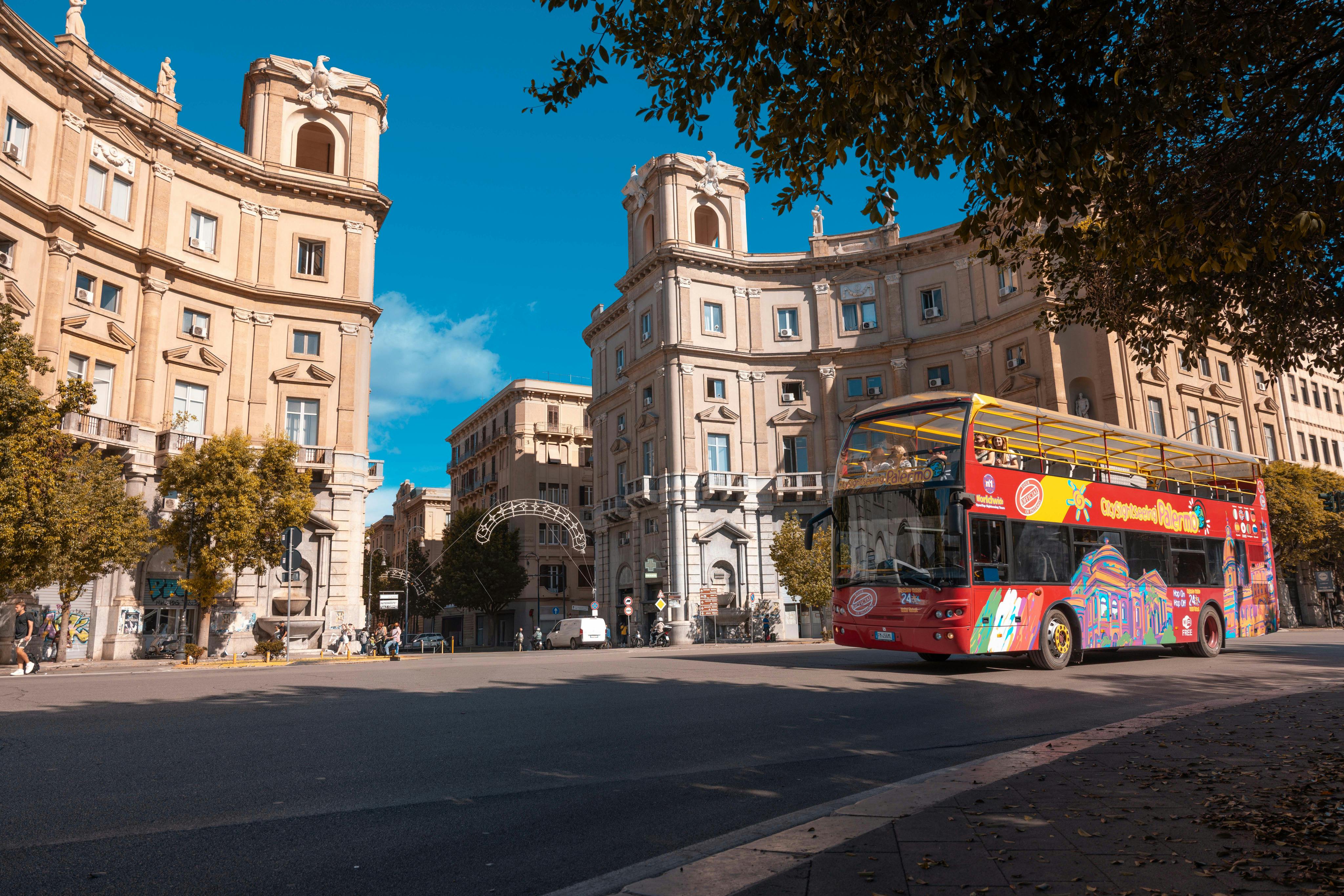 Un autobus turistico rosso a due piani su una strada soleggiata circondata da edifici storici e alberi.