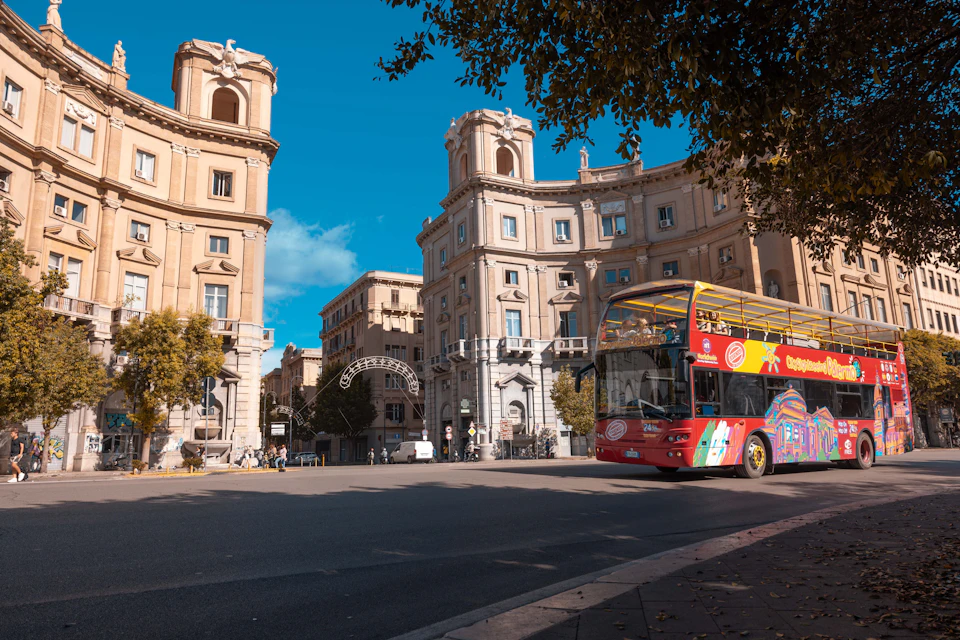 A red double-decker tour bus on a sunny street surrounded by historic buildings and trees.
