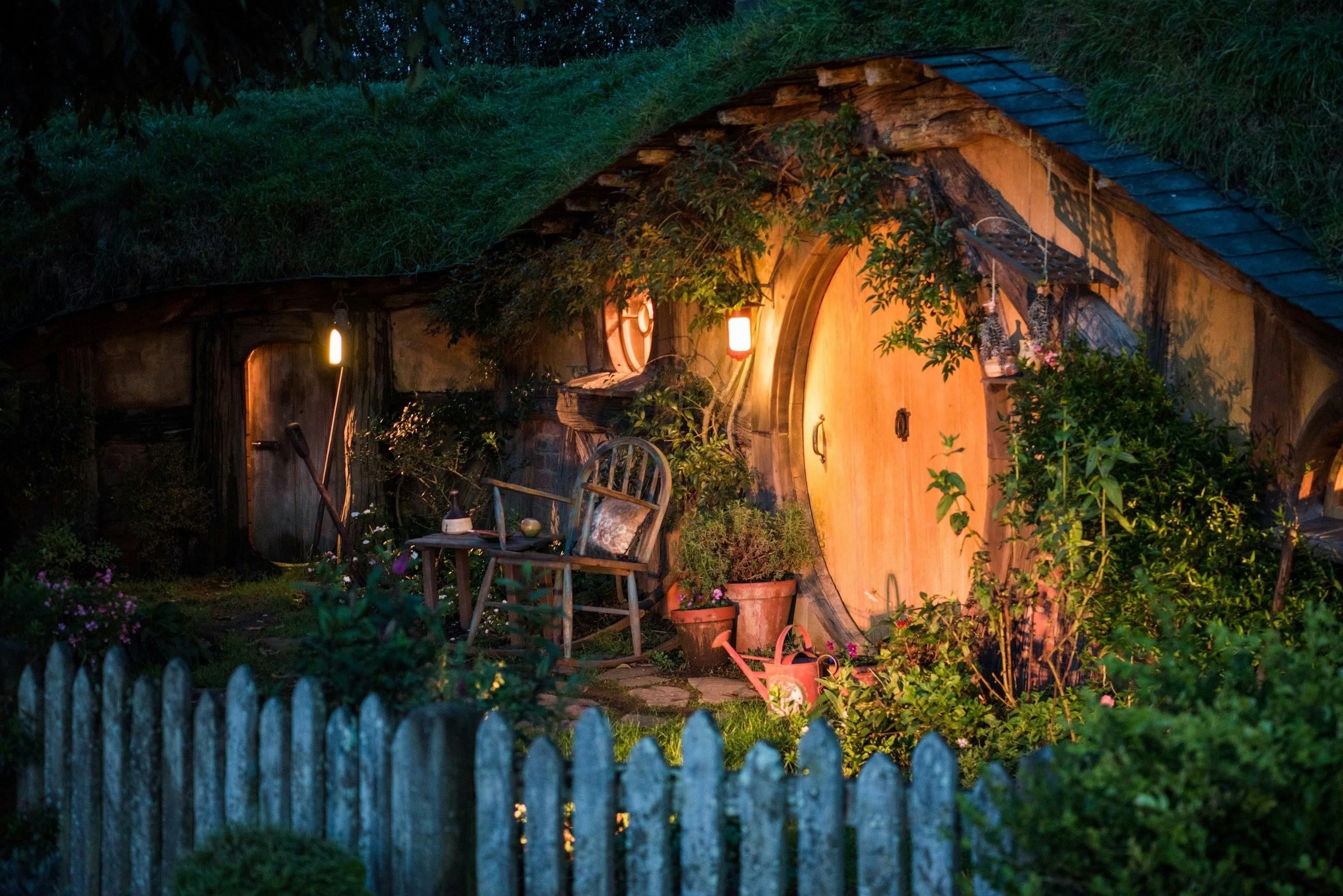 A cozy hobbit-style house with a round door, lit lanterns, and a rustic garden, surrounded by greenery and a wooden fence.