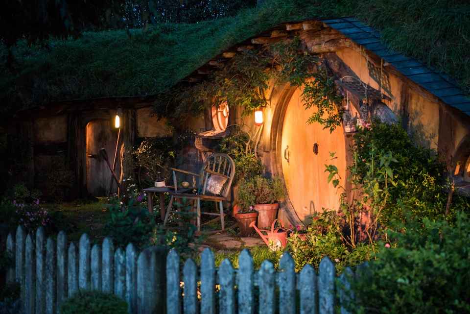A cozy hobbit-style house with a round door, lit lanterns, and a rustic garden, surrounded by greenery and a wooden fence.