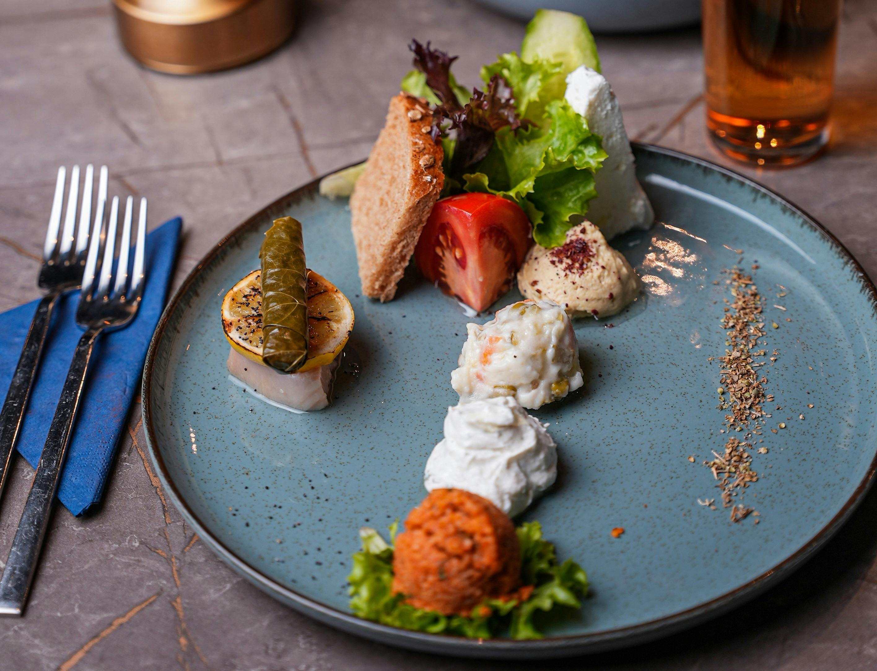 メッキした前菜には、ドルマ、フムス、ツァツキ、サラダ、パン、トマトのスライスなど、さまざまな小さなサービングが青い皿に盛られています。